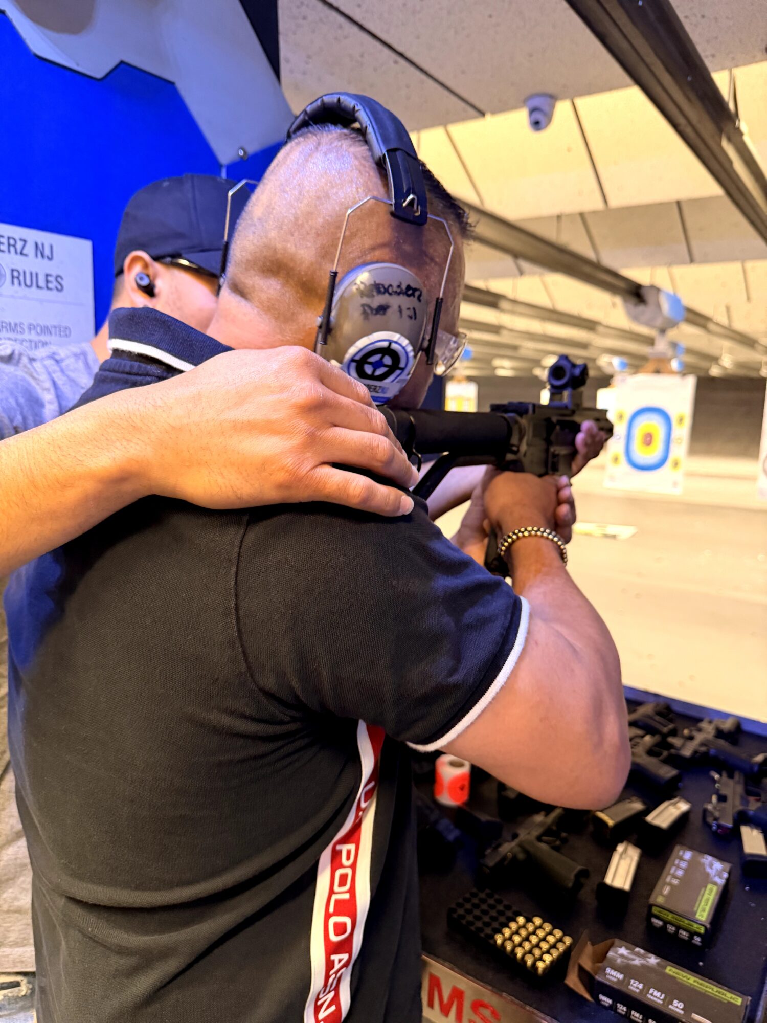 Person at shooting range aiming a firearm, wearing ear protection and a black shirt.