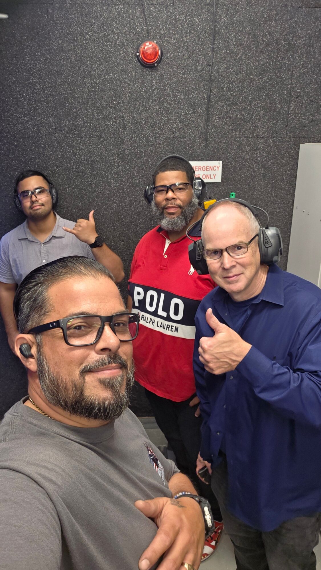 Four people in an elevator, smiling and taking a selfie, with a gray wall and a red fire alarm in the background.