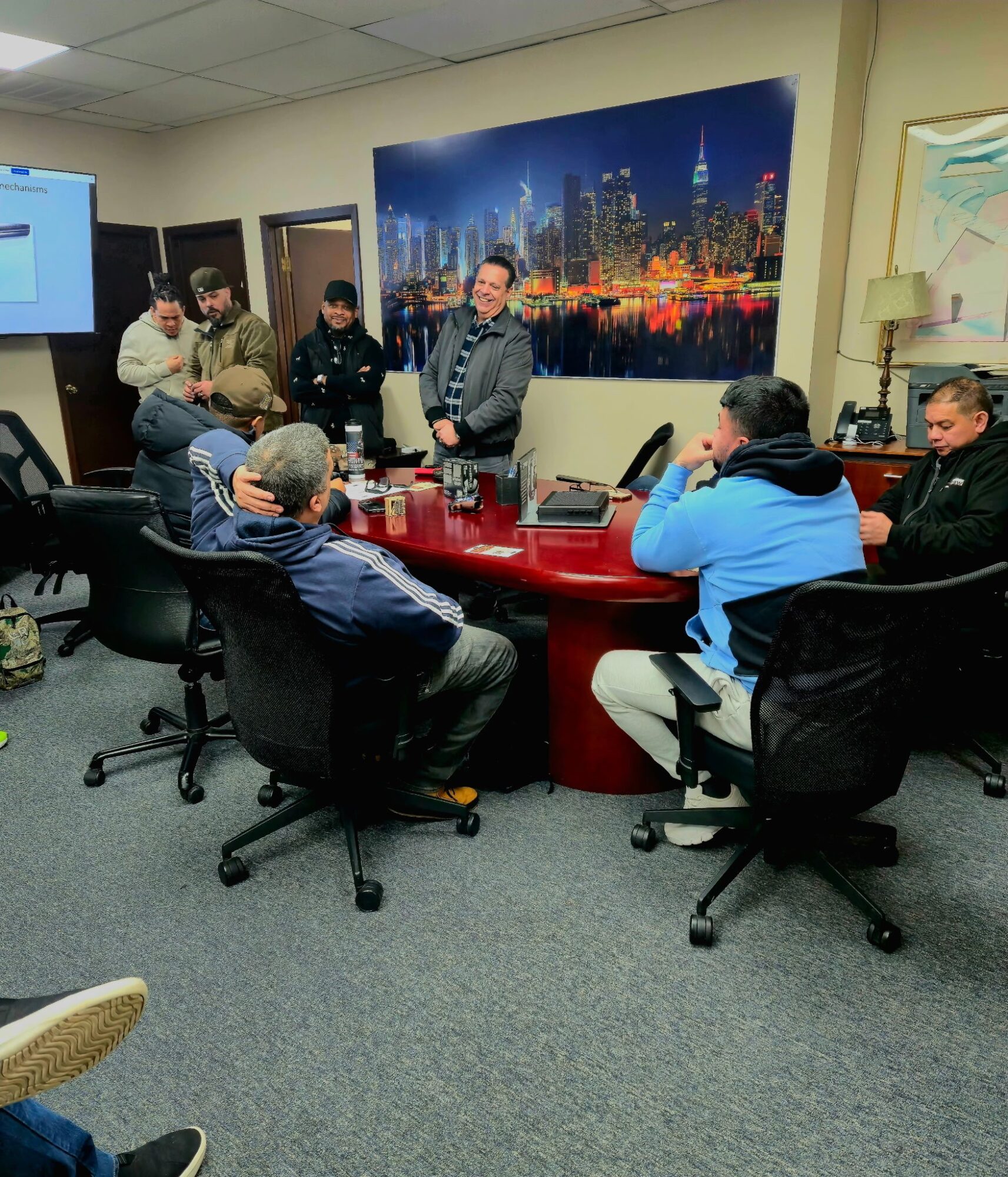 Group of people in a meeting room around a large red table, with a city skyline picture on the wall.