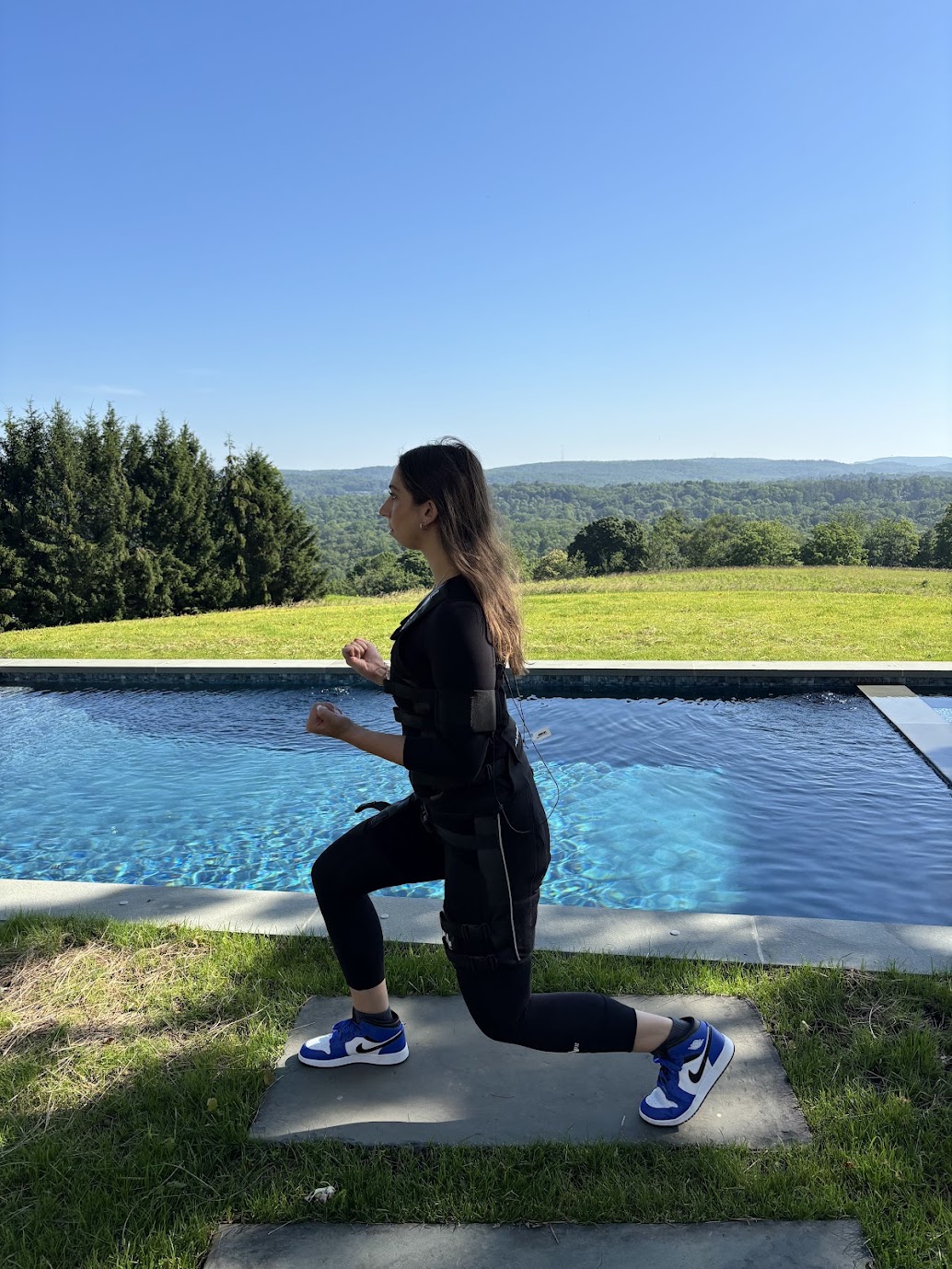 Woman in black workout clothes and sneakers kneeling on a mat by a pool outdoors, with a landscape of trees and hills in the background.