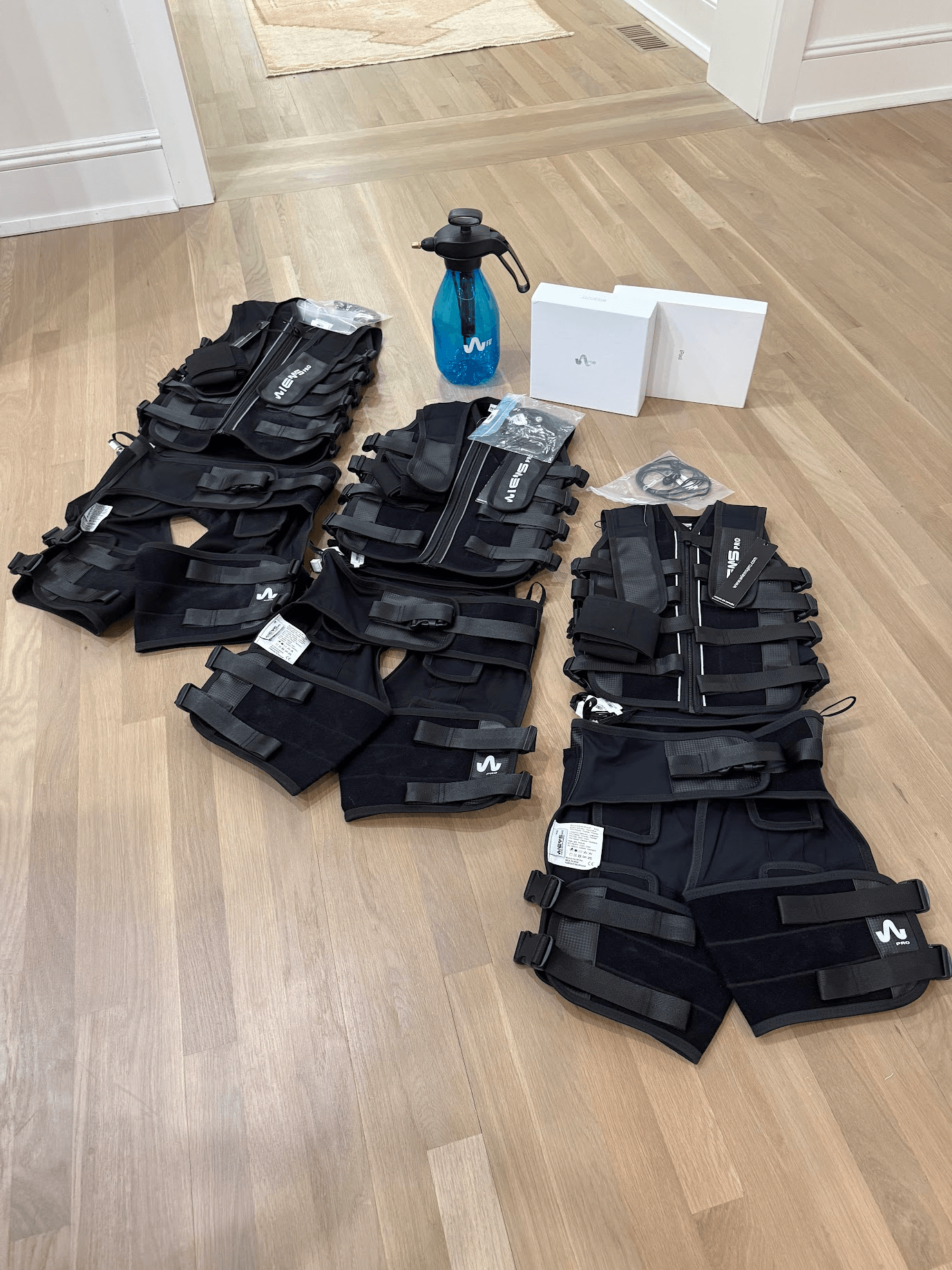 Multiple black tactical vests, a blue spray bottle, and white boxes on a wooden floor.