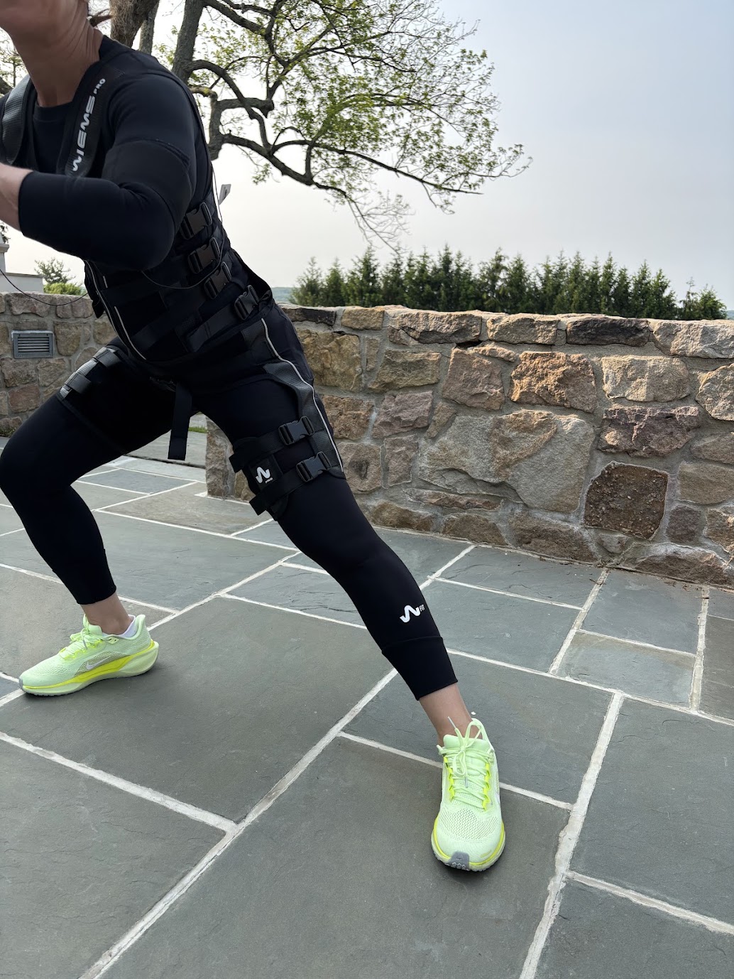 Person in black athletic clothing and neon yellow shoes stretching outdoors on tiled ground with stone wall and trees in background.