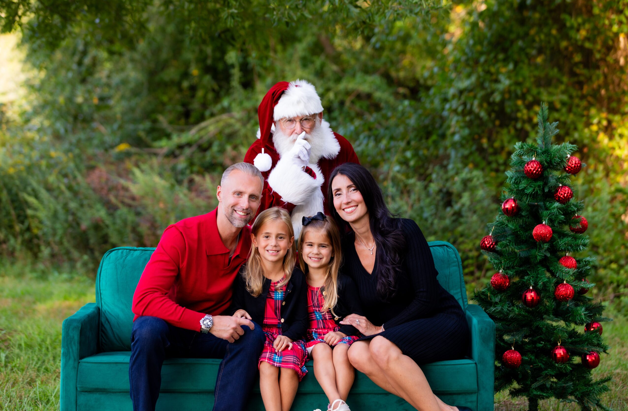 Family sitting on a green sofa outdoors with a person dressed as Santa Claus, a decorated Christmas tree, and trees in background.