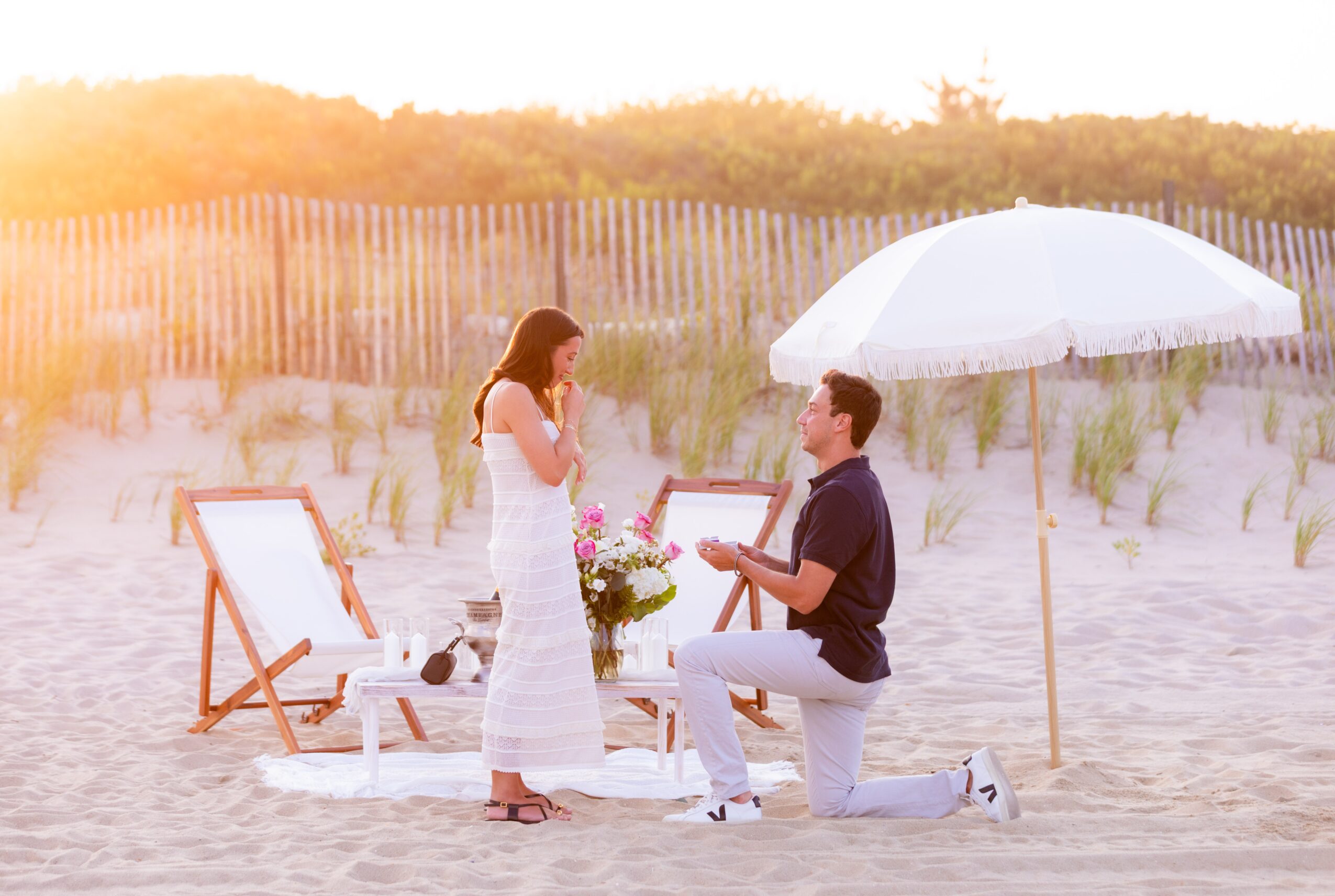 A man proposes to a woman on a beach with a bouquet, under a white umbrella, with a chair and fence in background.
