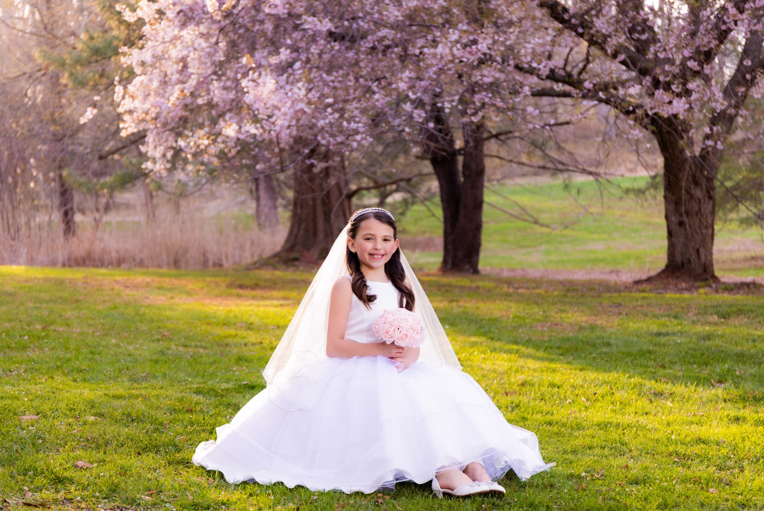 Young girl in a white dress and veil sitting on grass with pink flowering trees in background.