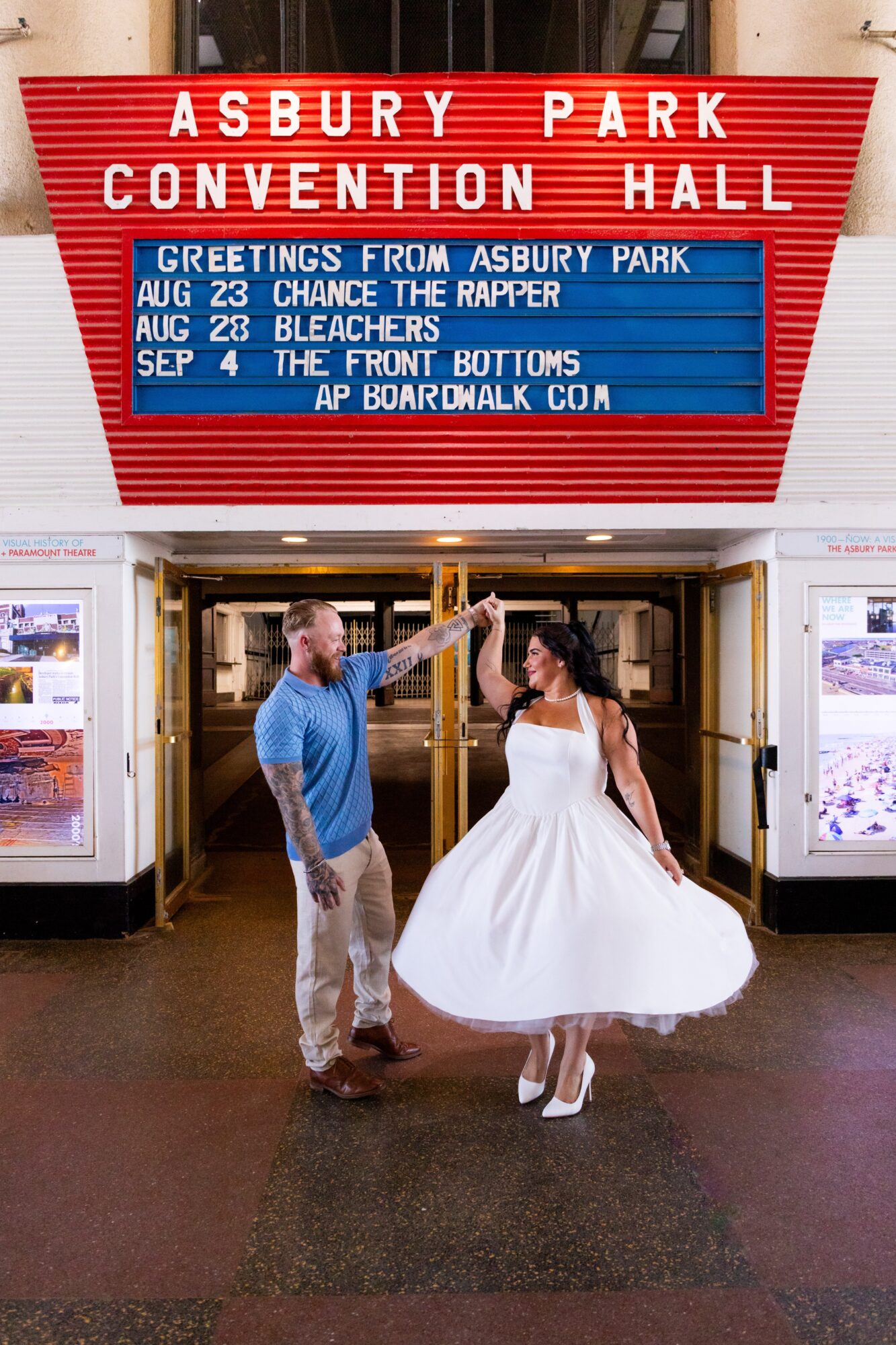 Two people dance in front of a theater entrance with a marquee displaying event information, one in a blue shirt and the other in a white dress.