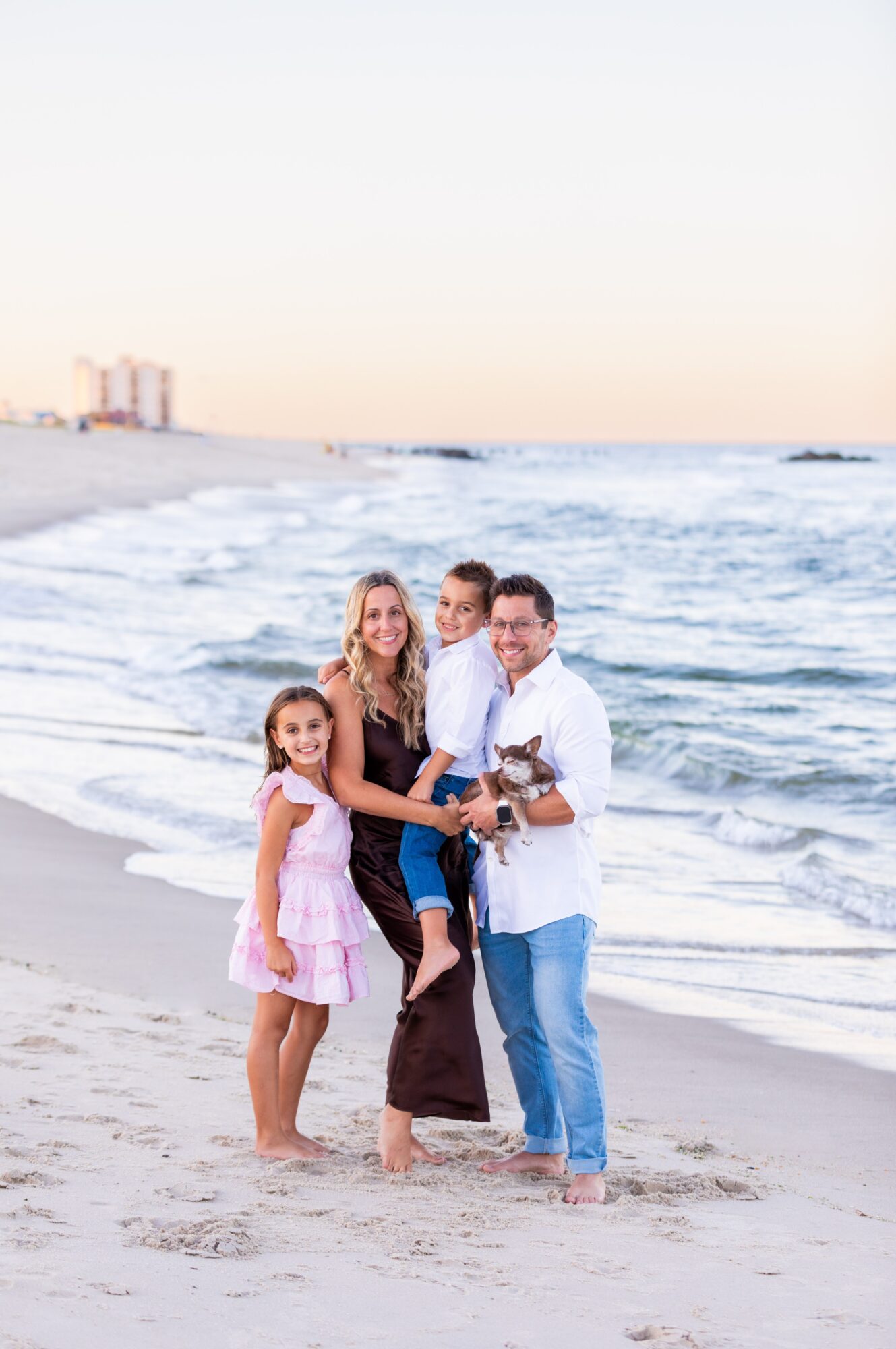 Group of four people standing on a beach with ocean waves in the background, smiling and holding a small dog.