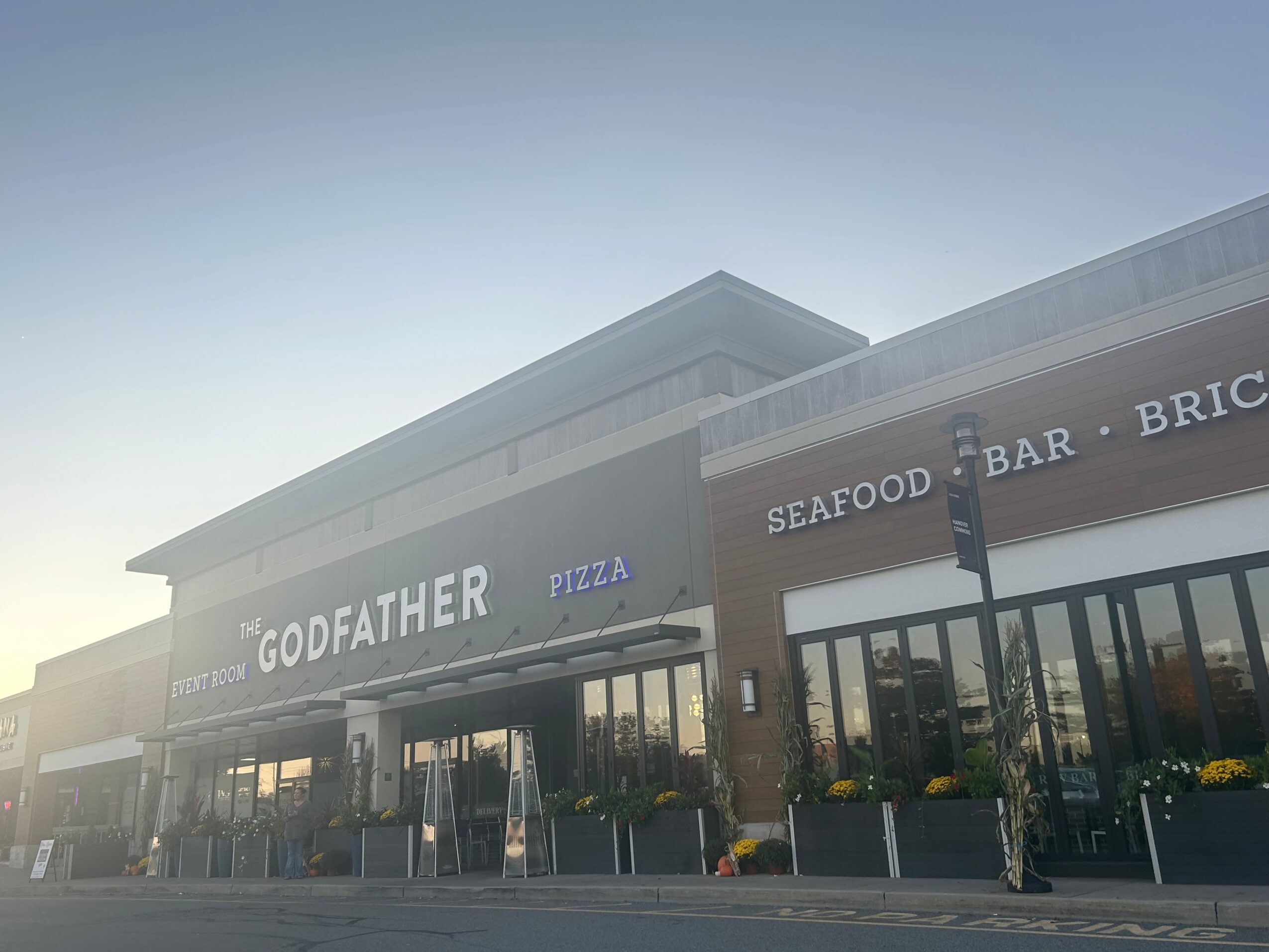 Exterior view of a restaurant with large windows, signage for seafood, bar, pizza, and Godfather, with outdoor seating.