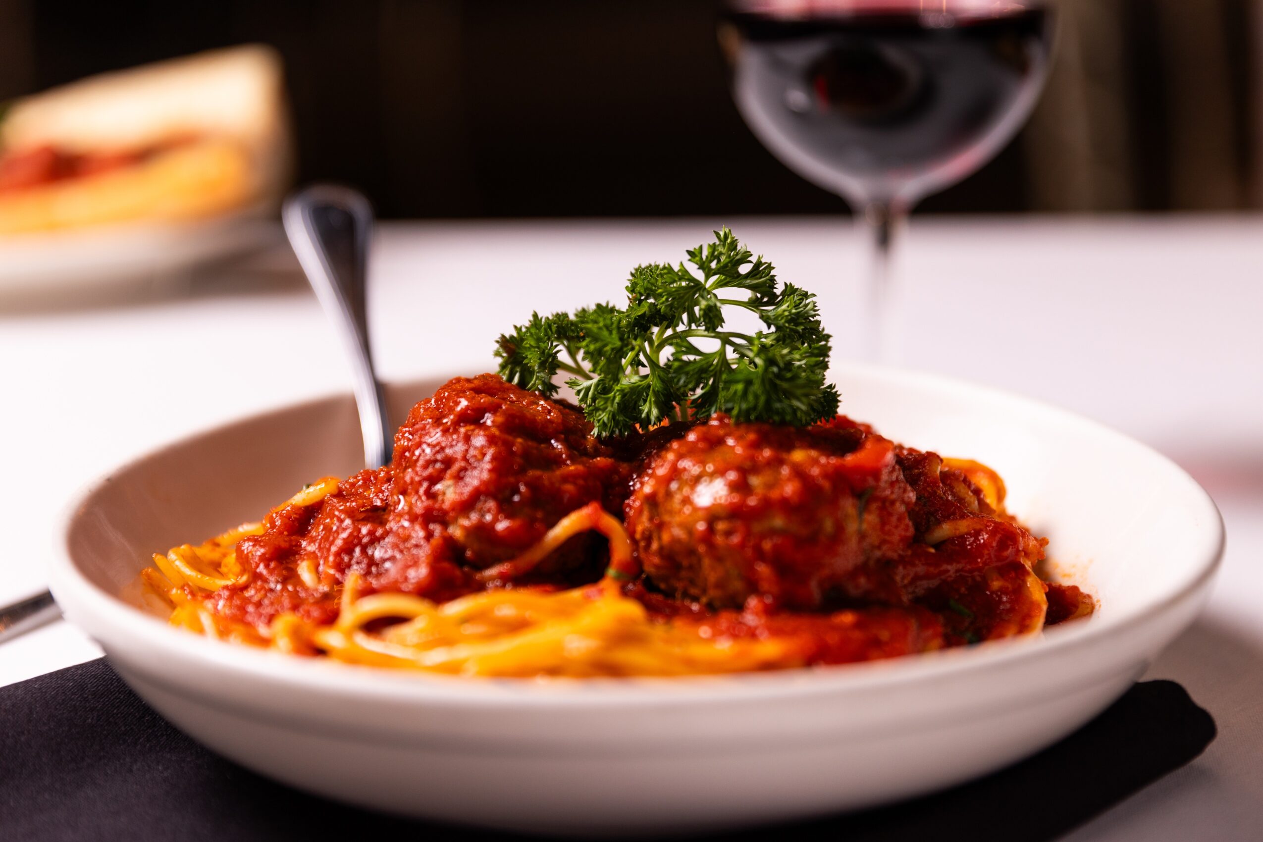 Plate of spaghetti with meatballs, garnished with parsley, with a glass of red wine in the background.