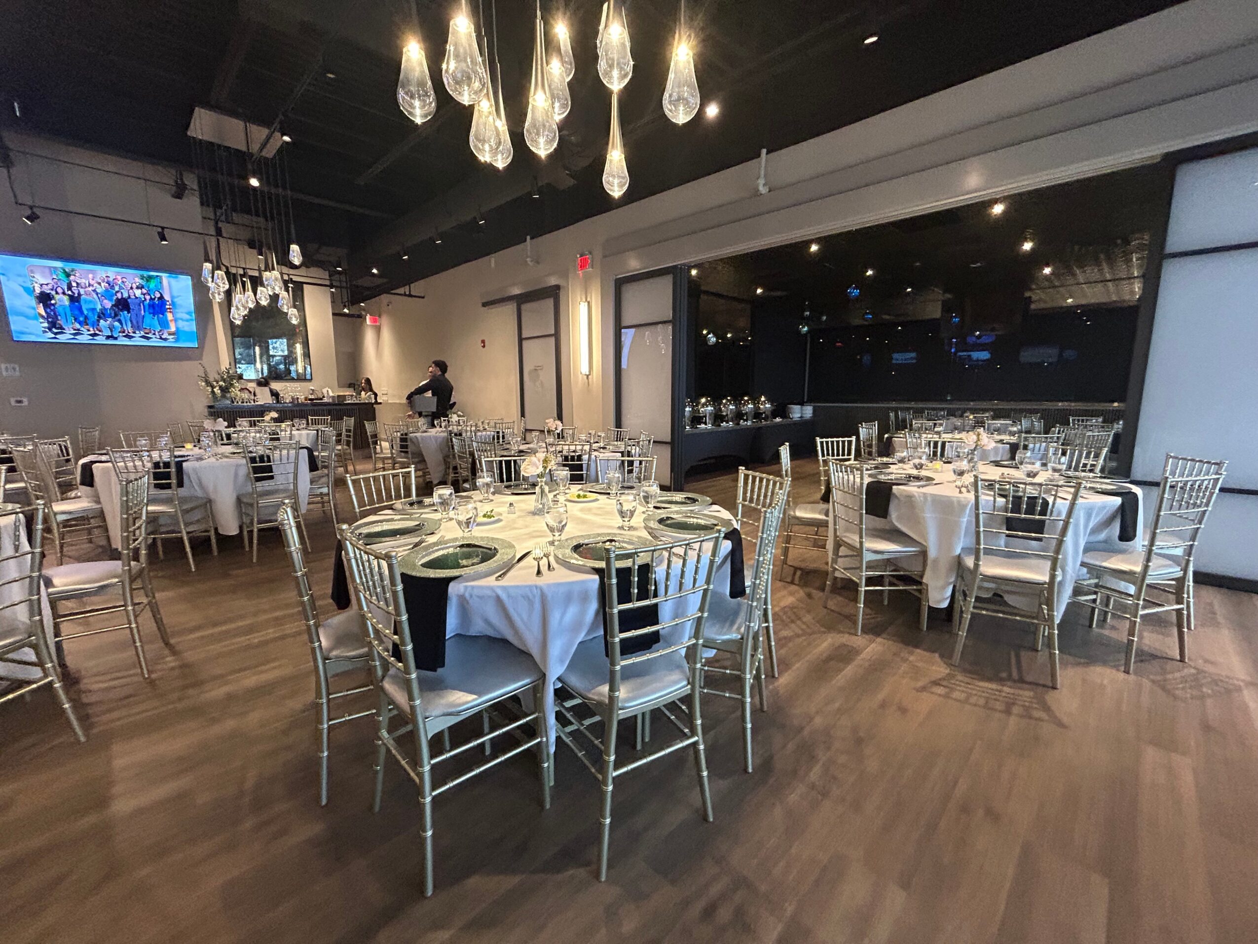 Elegant dining area with round tables, white tablecloths, and silver chairs, illuminated by hanging pendant lights, with a large window view.
