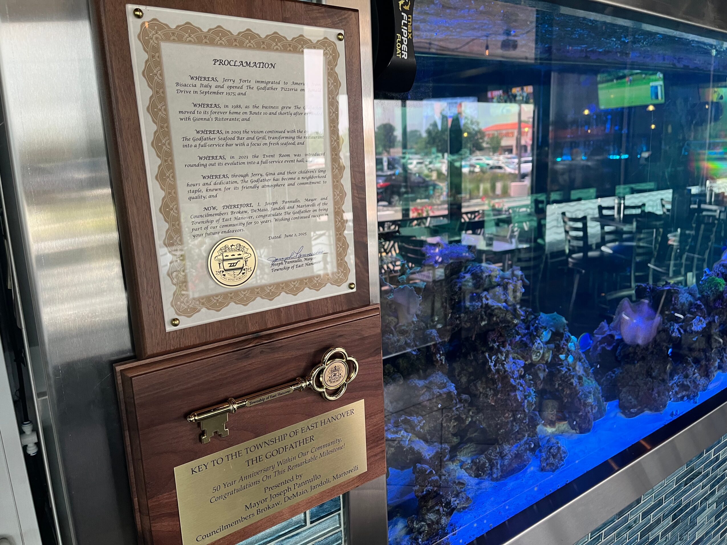Framed document and a brass plaque on a wooden wall, with a glass window showing a restaurant interior with tables and chairs, and a blue-lit aquarium.