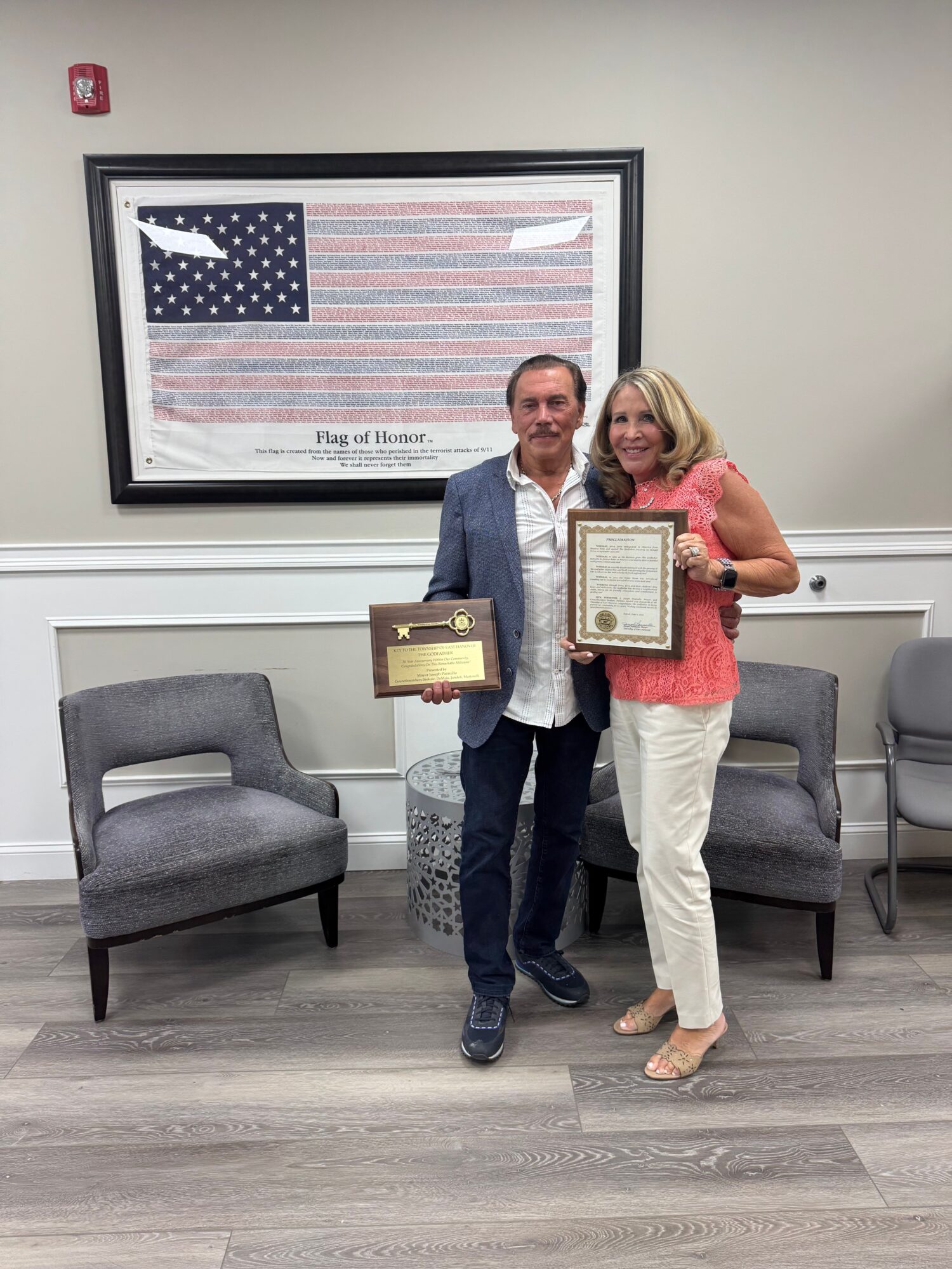 Two people standing in a room with chairs and a framed American flag on the wall, holding certificates or awards.