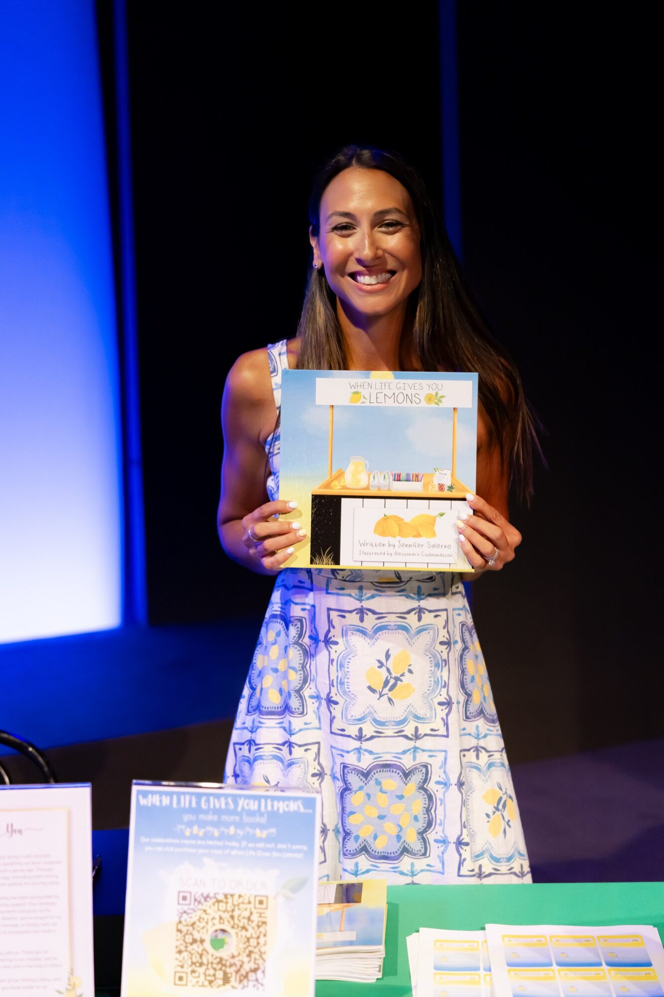 Smiling woman holding a colorful certificate on stage, wearing a patterned dress, with awards and books on table nearby.