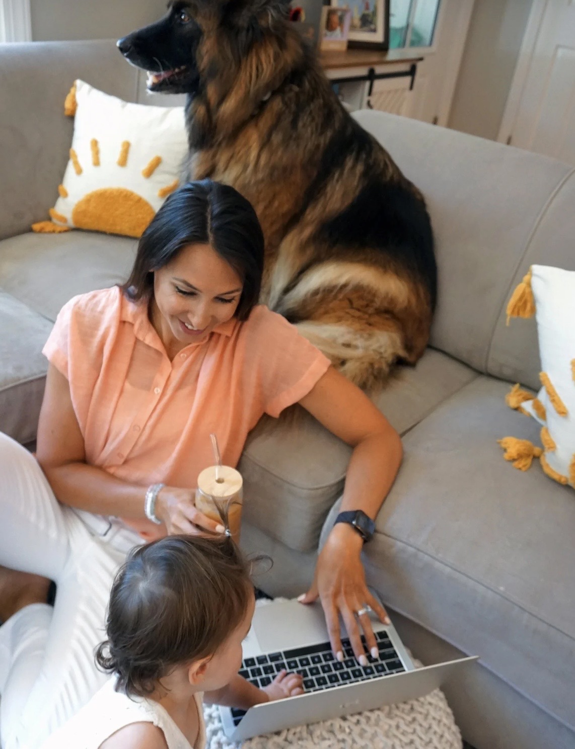 Woman and child sitting on a sofa with a large dog behind them, using a laptop and holding a drink, in a cozy room.