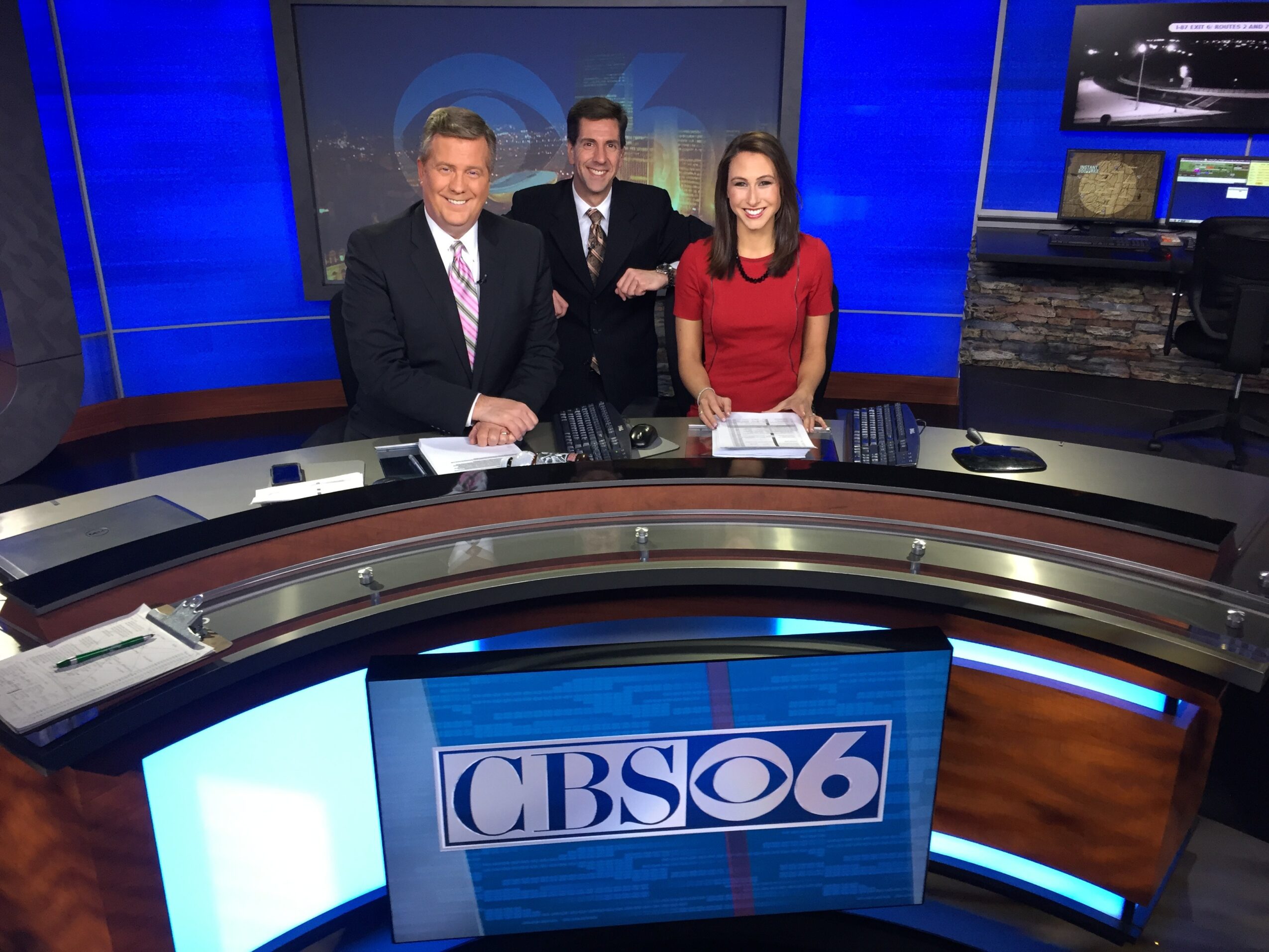 Three news anchors sit behind a curved desk with a CBS6 logo, in a television studio with blue background and monitors.