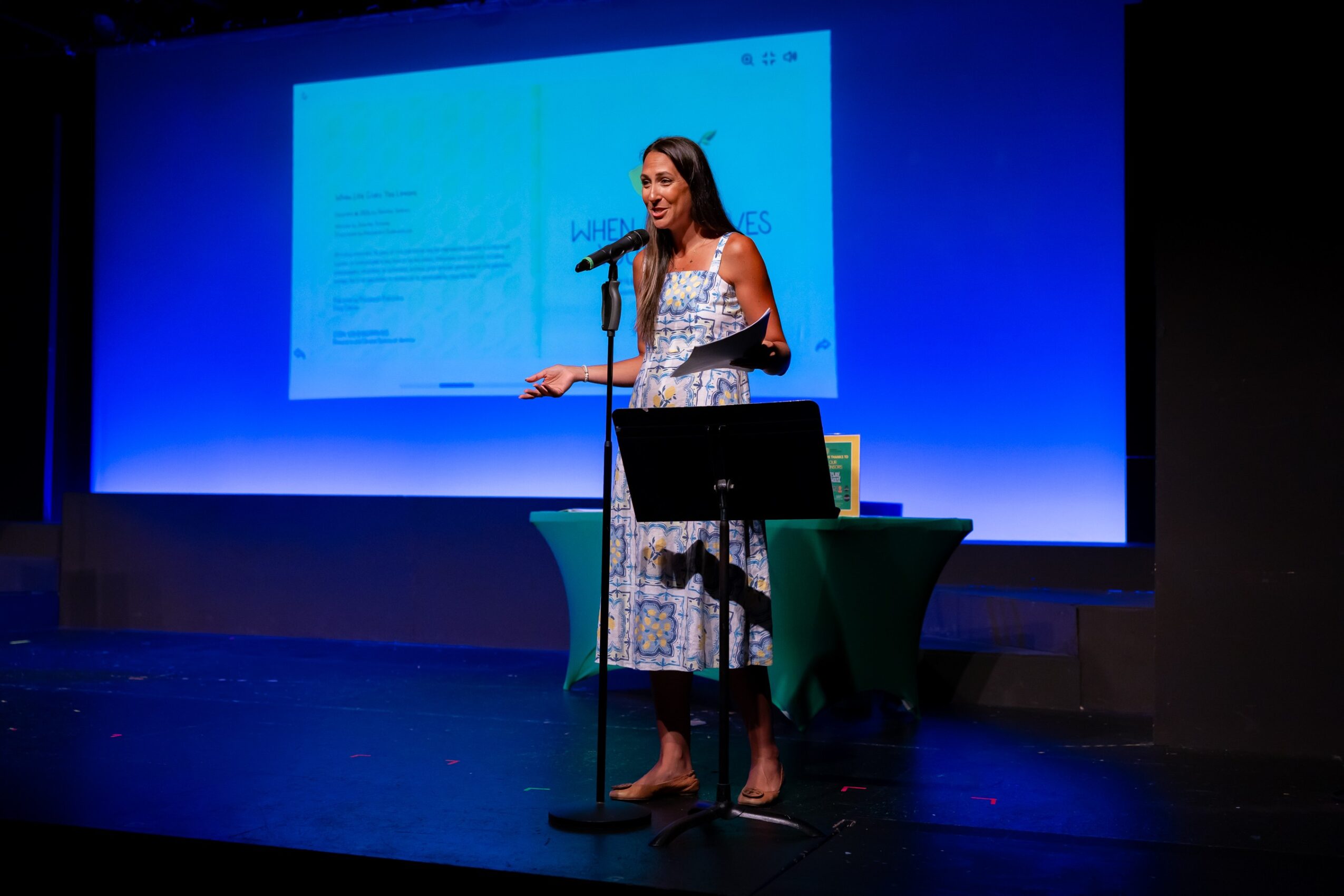 Woman speaking at a microphone on stage with a presentation slide behind her, standing next to a small table with a book and a glass.
