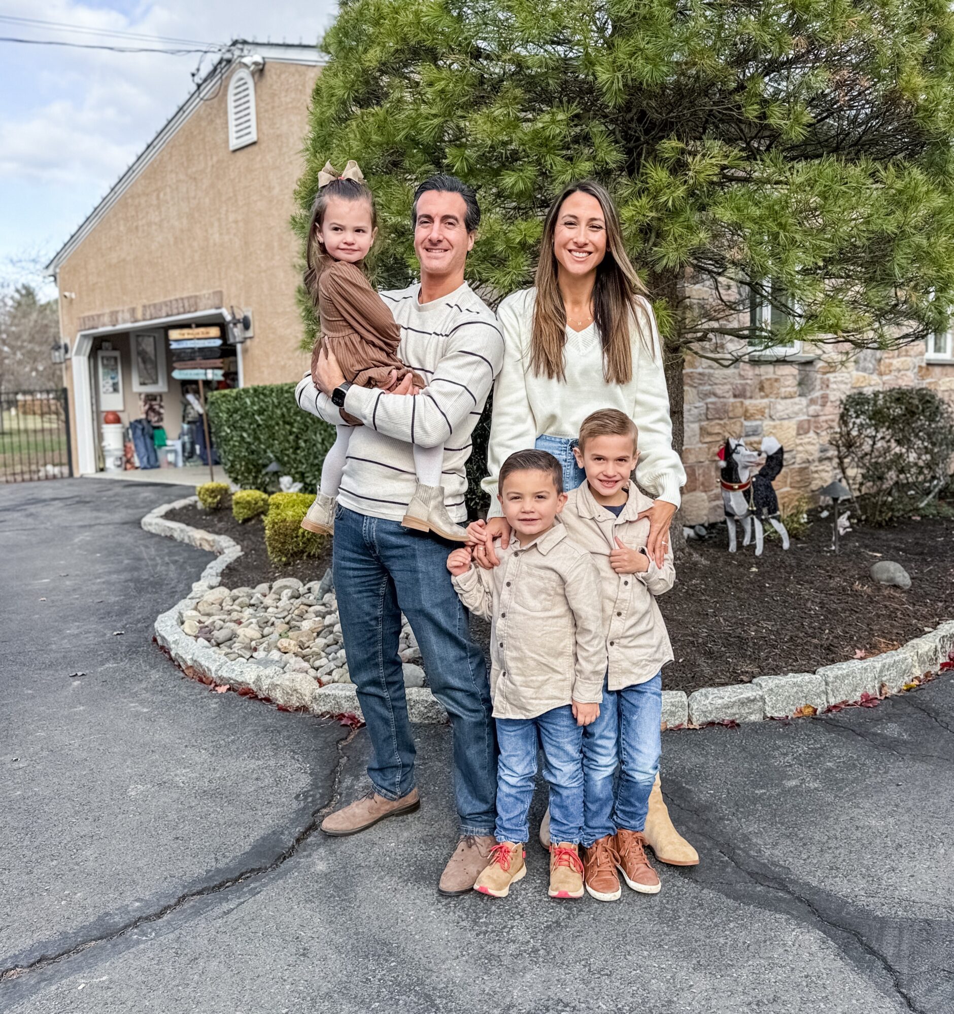 Family of five standing outdoors on a paved area with a building, trees, and a motorcycle in the background.