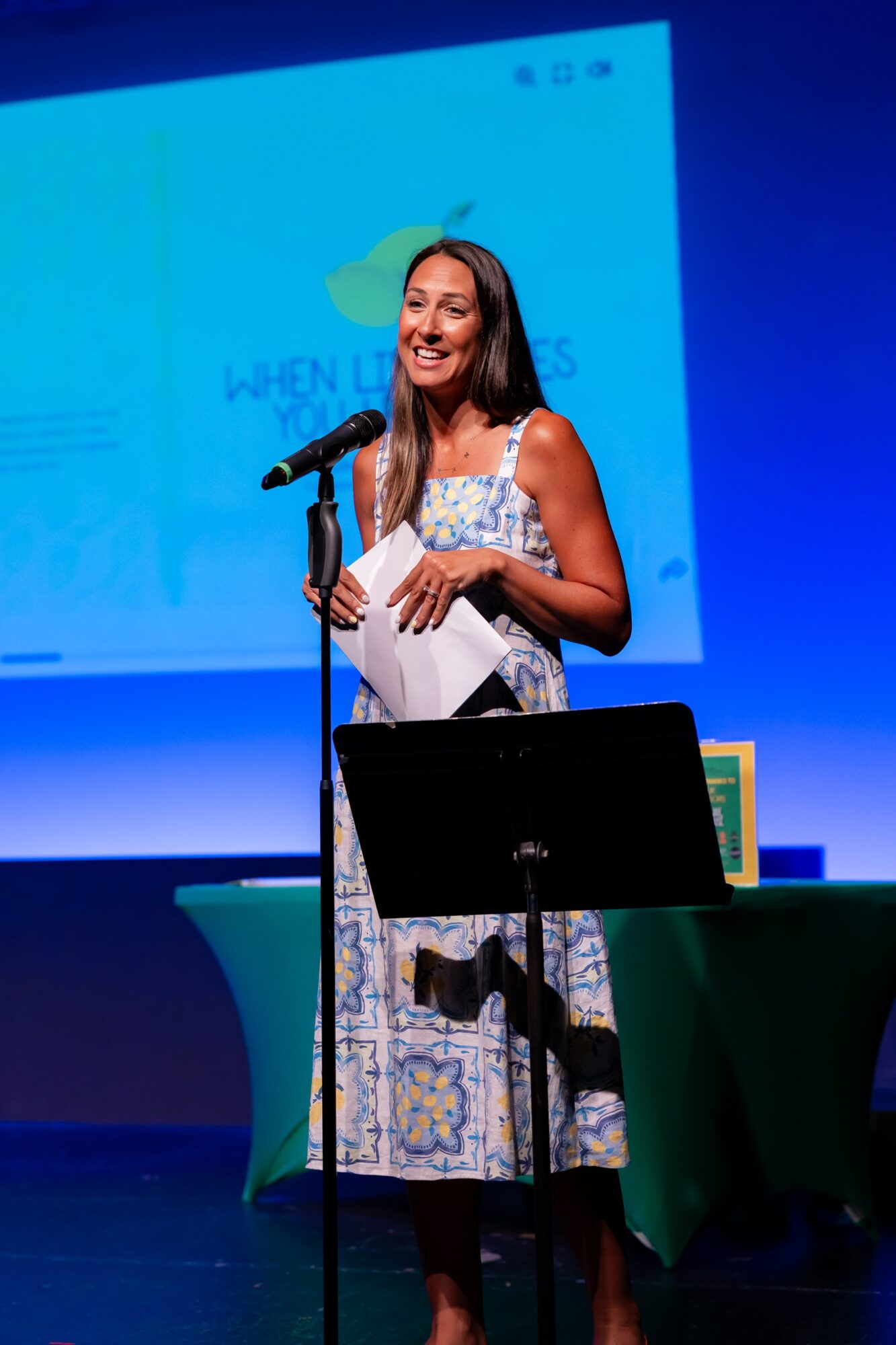 Woman standing on stage, smiling, holding papers, with a microphone, in front of a blue screen.