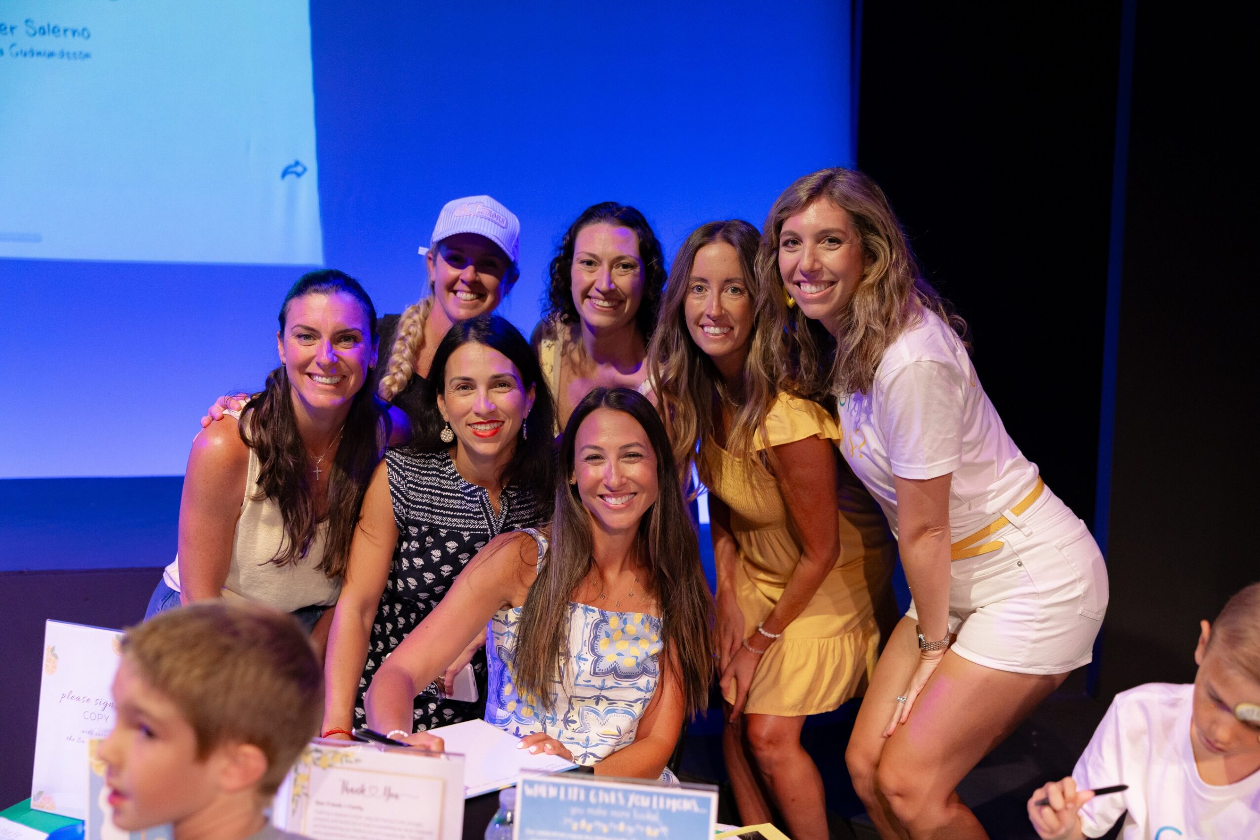 Group of eight women and one child smiling on stage, some wearing casual summer clothing, with a blue background and a screen.