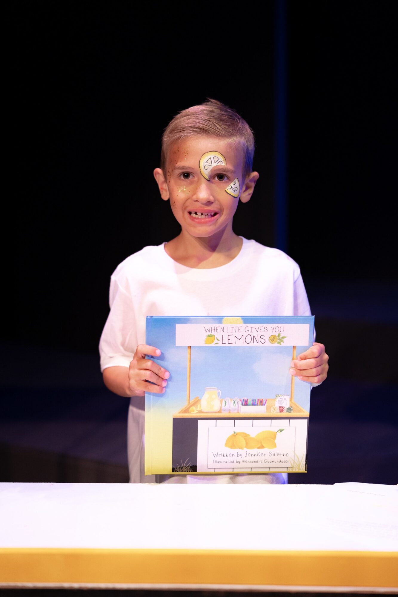 Child with face paint holding a colorful box, smiling, against a dark background.