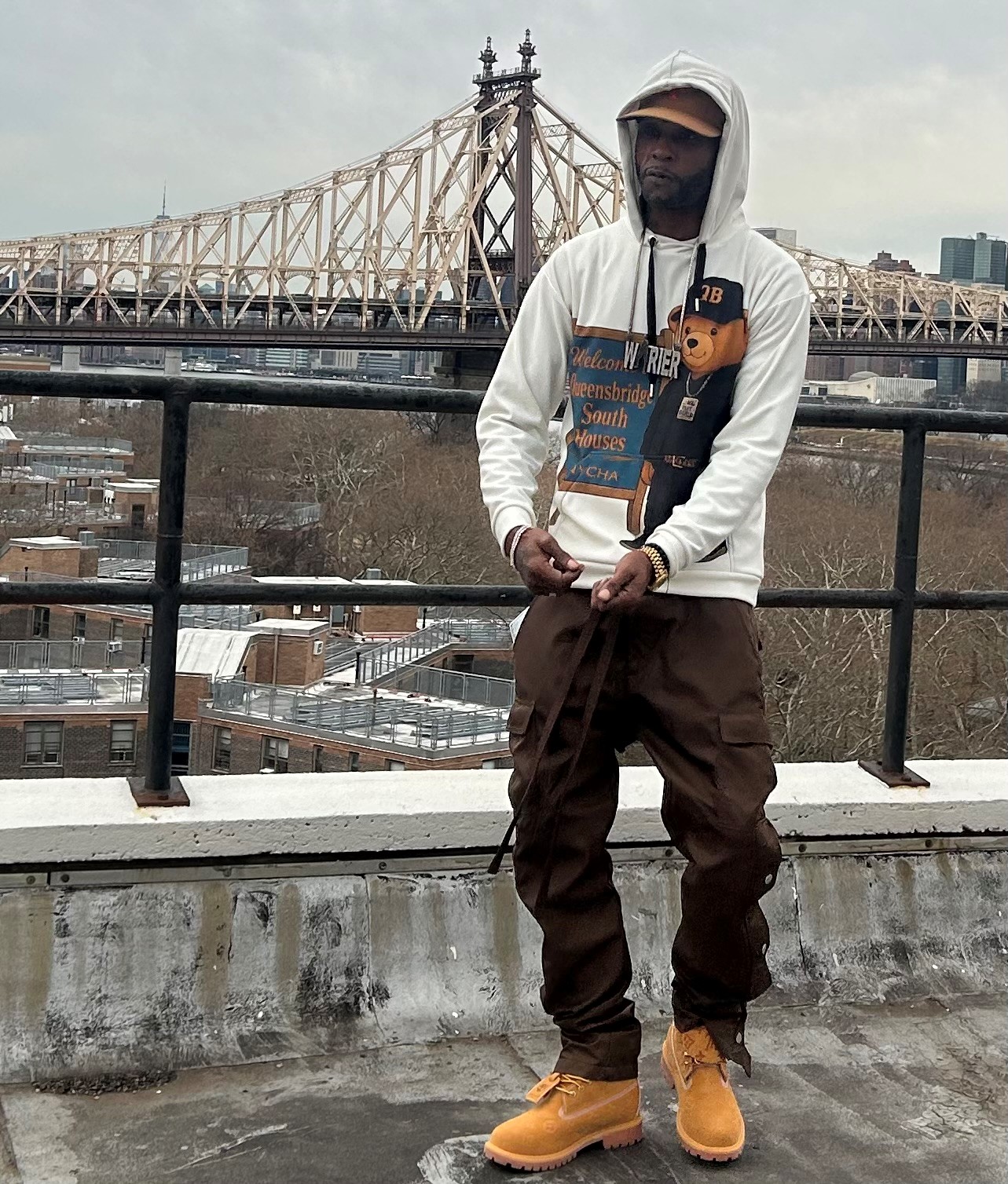 Person standing on rooftop with cityscape and bridge in background, wearing hoodie, sunglasses, and tan boots.