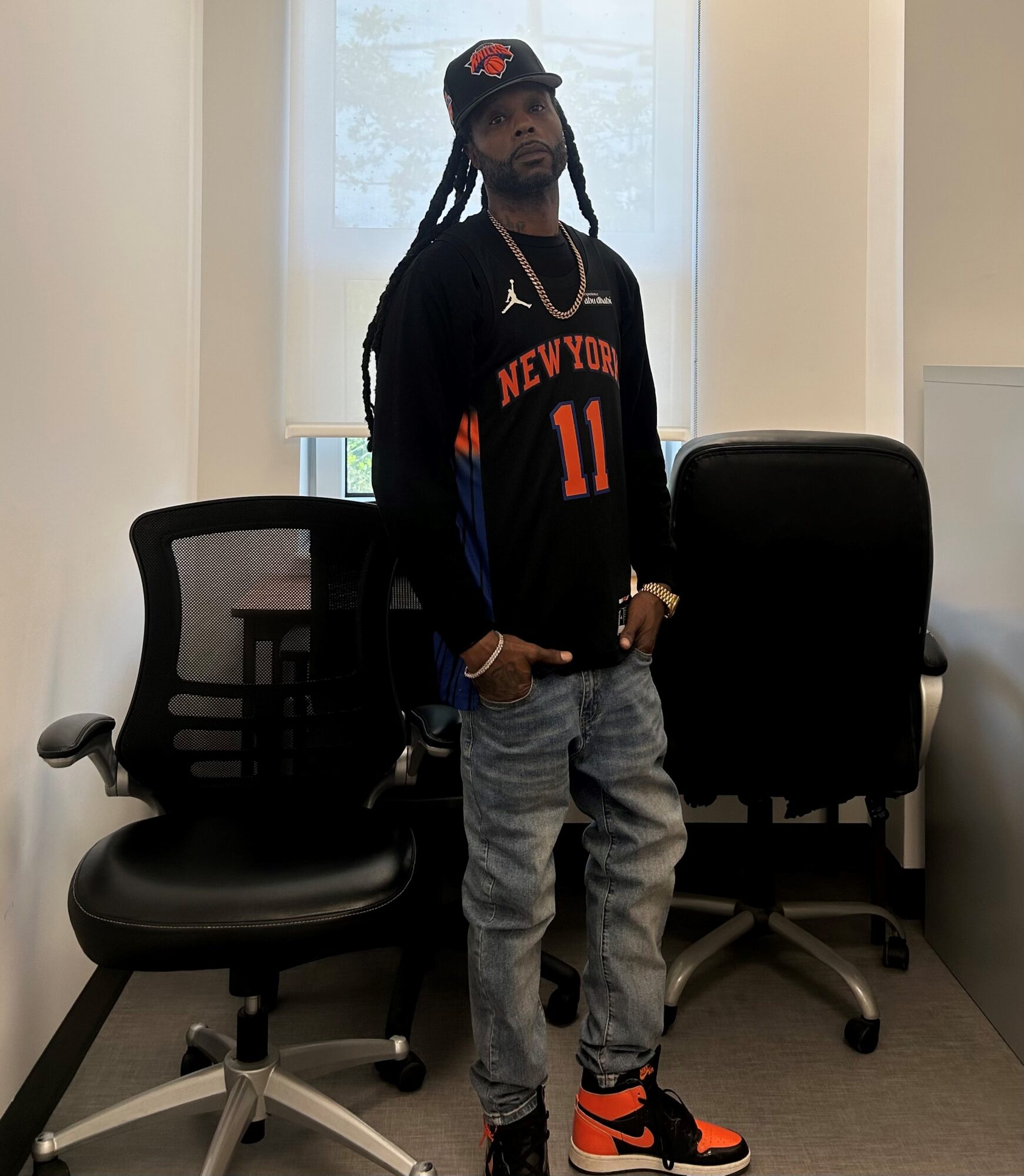 Young man with dreadlocks wearing a black New York basketball jersey, jeans, and sneakers, standing in an office room.