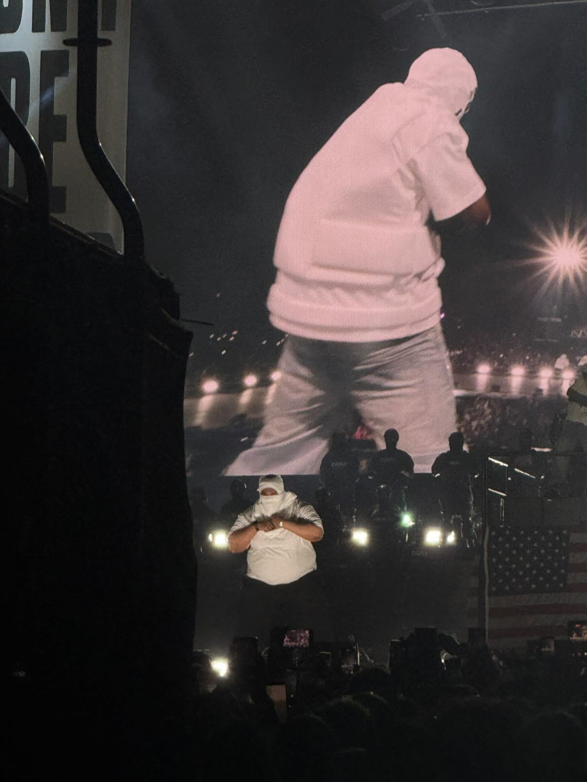 Person on stage with large screen behind, holding a microphone, wearing a light-colored shirt and pants, performing at a concert.