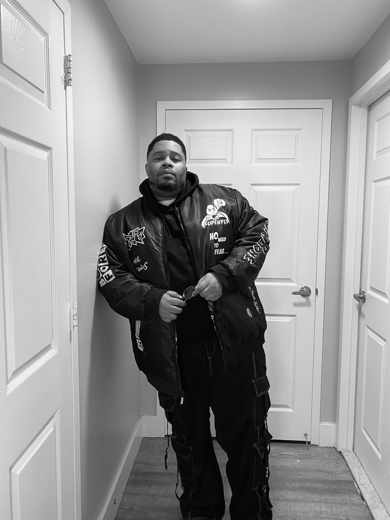 Person standing in a hallway, wearing a black jacket with patches, holding a bag, with closed white doors behind.