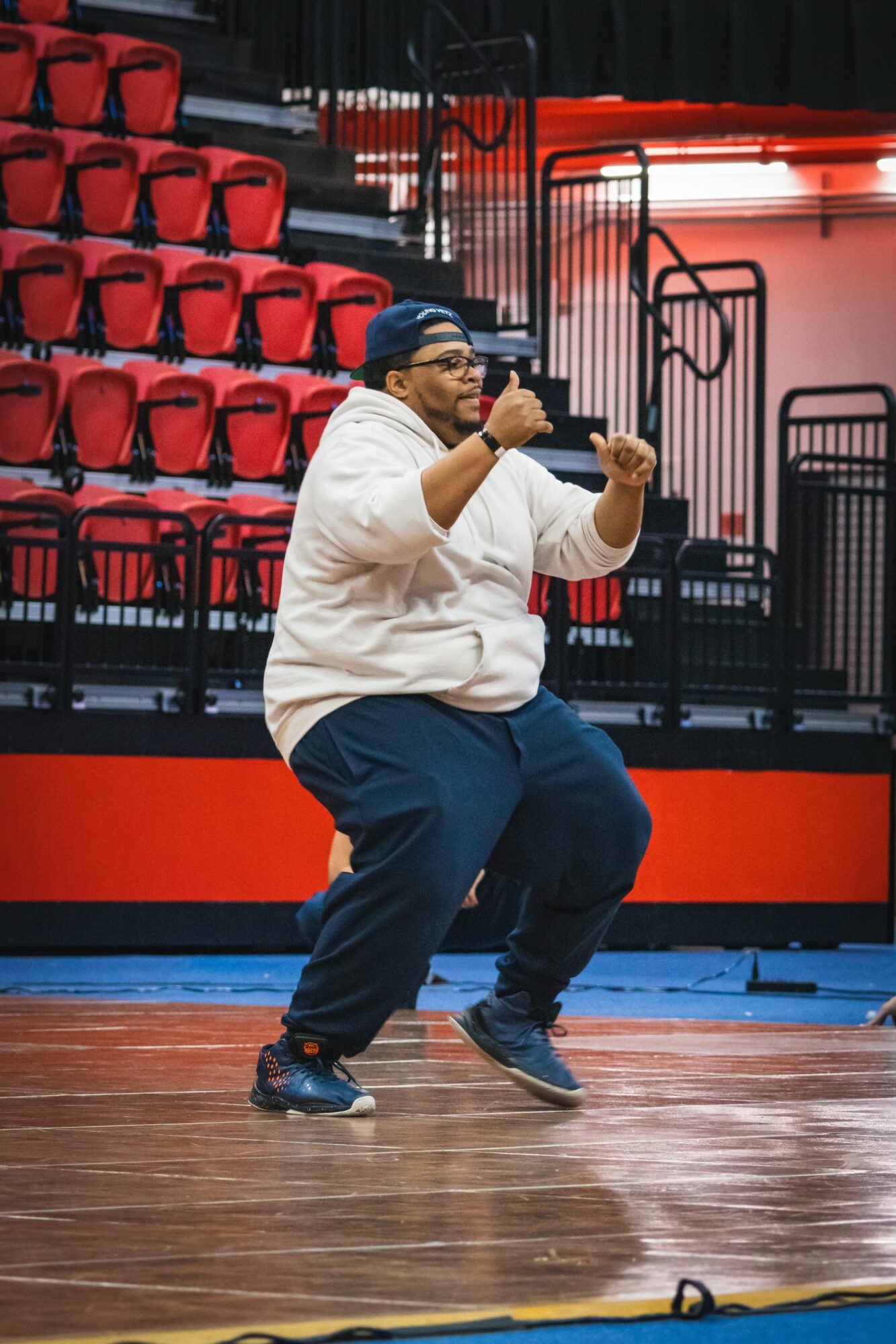 Person dancing on a basketball court with red stadium seats in the background, wearing a white hoodie, blue pants, and a cap.