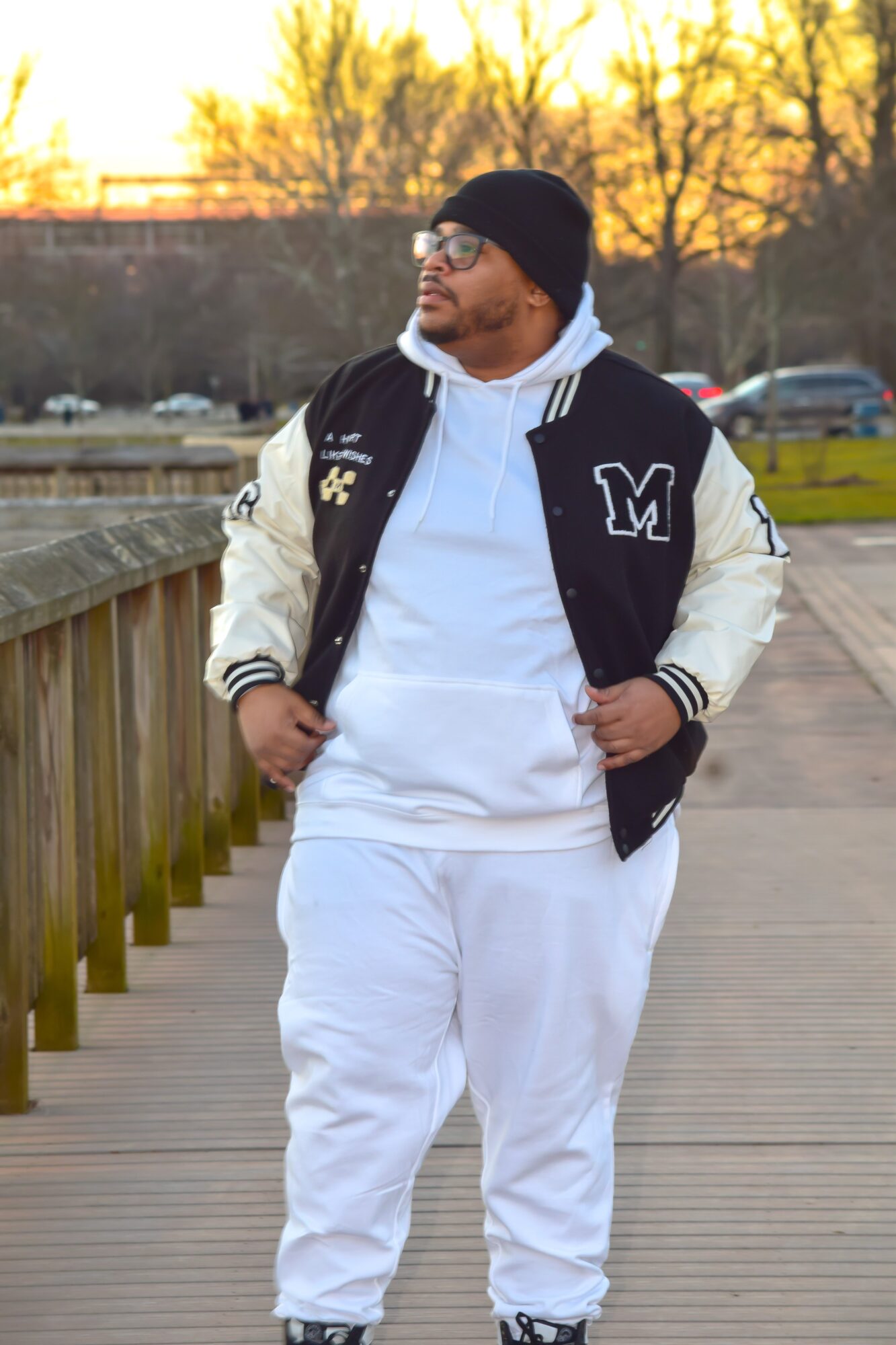 Man walking on a wooden pathway wearing a black and white varsity jacket, white hoodie, white pants, and a black beanie, with trees and cars in the background.