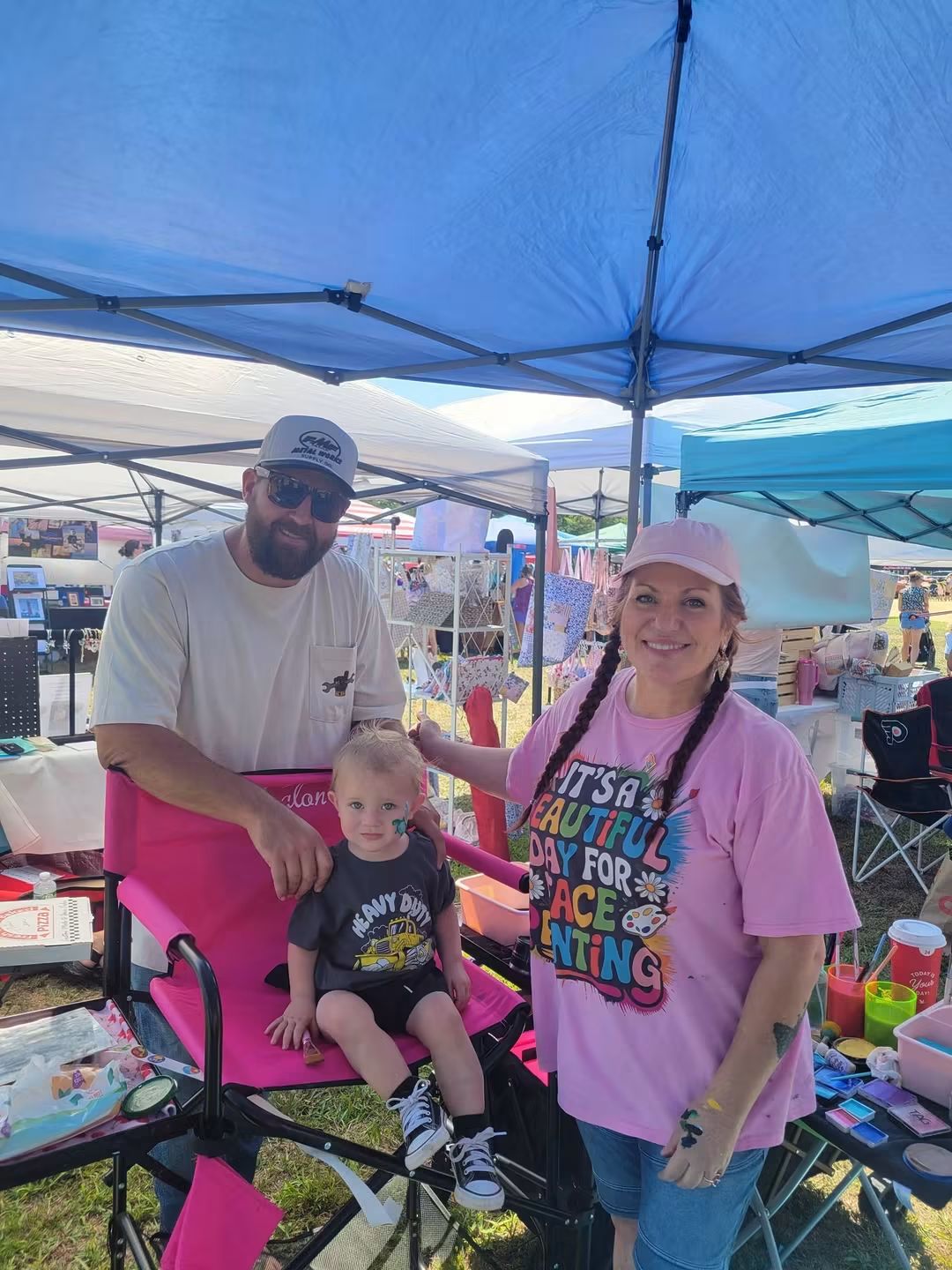 A man, a young child, and a woman under a blue canopy at an outdoor event, with tables and tents in the background.