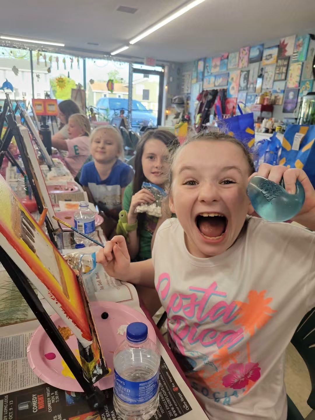 Children at a craft table in a classroom, smiling and excited, with art supplies and colorful decorations around.