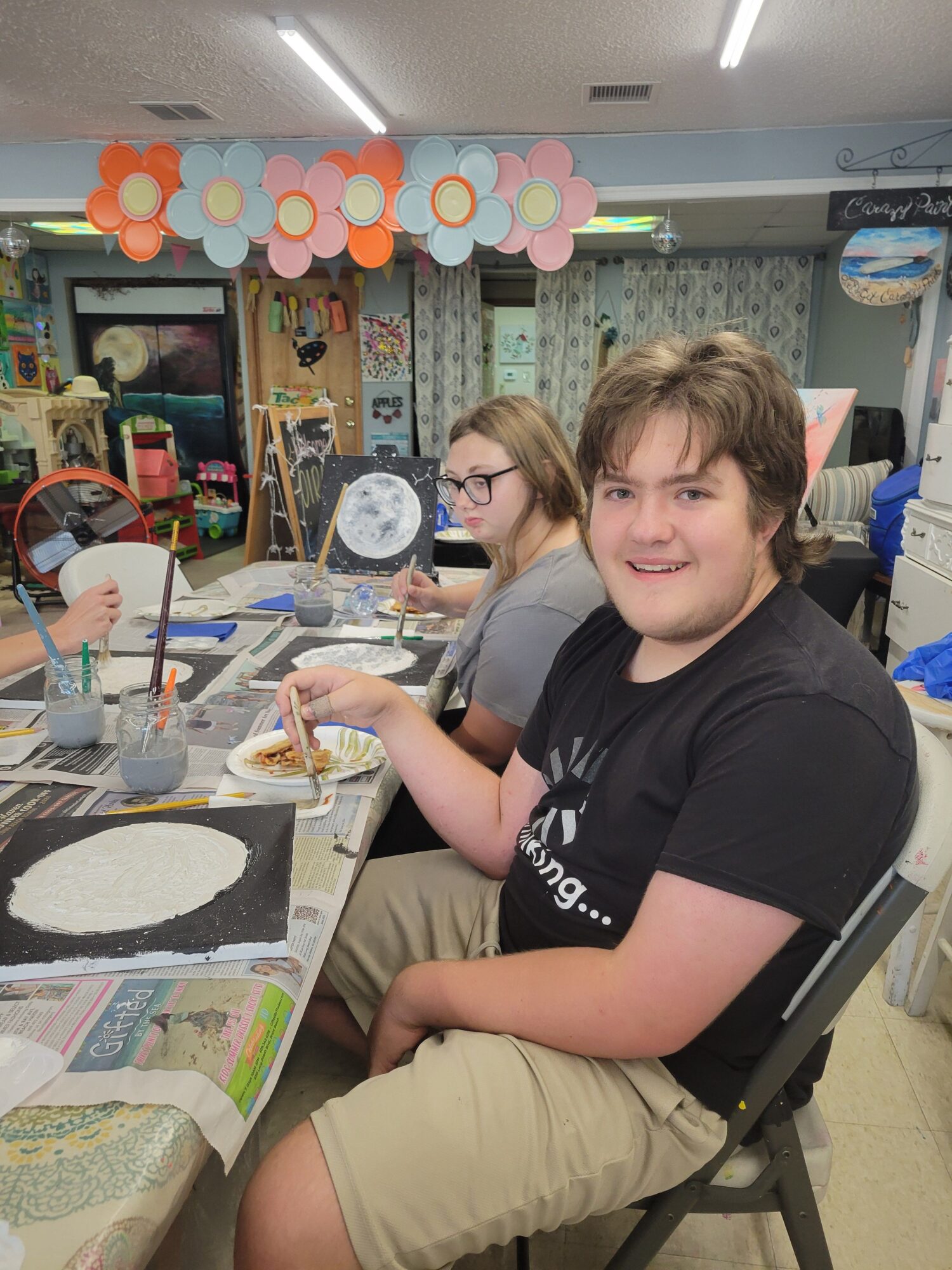 Two children sit at a table with food and drinks, smiling at the camera in a room with colorful decorations.