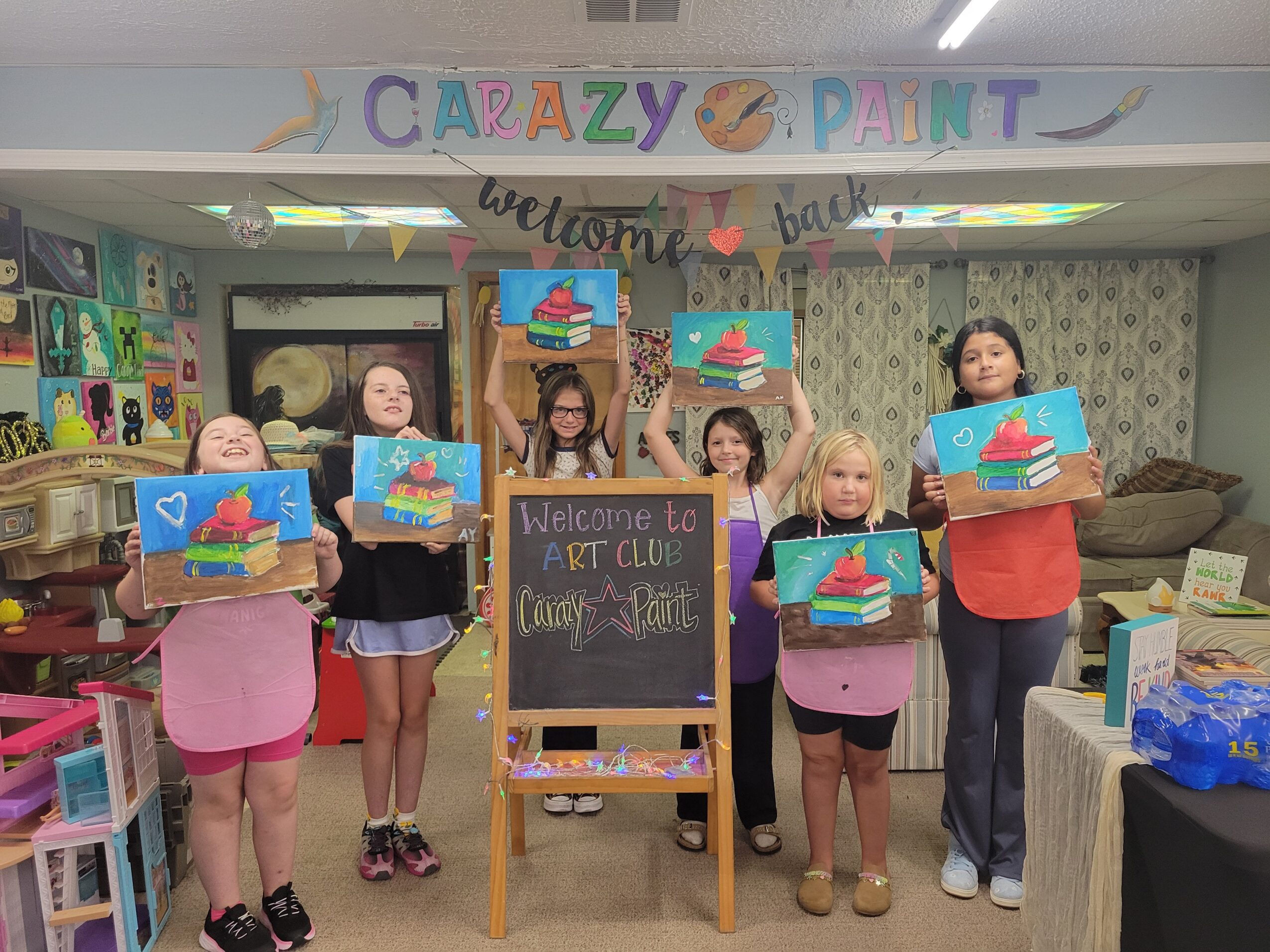 Seven children holding paintings in a colorful art studio with a chalkboard and decorations, celebrating a painting event.