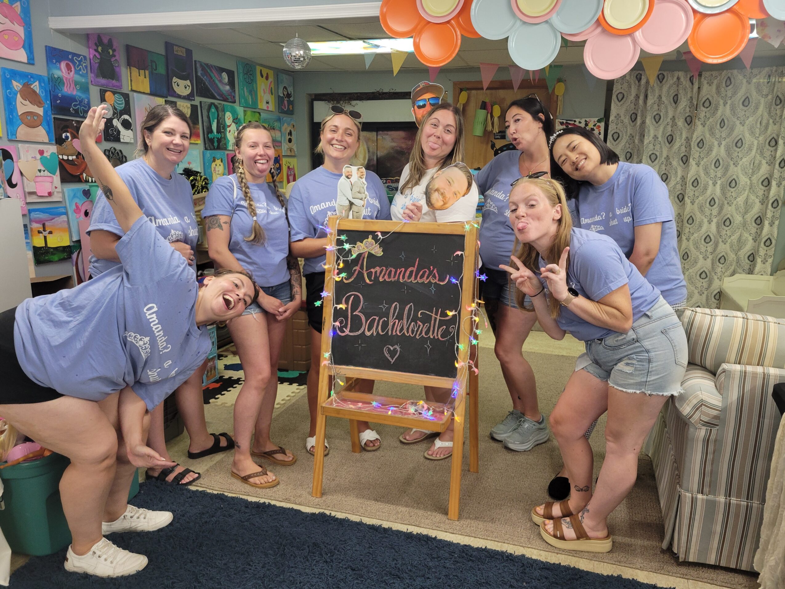 Group of eight young women in matching blue shirts gathered around a chalkboard sign in a decorated room, smiling and posing.