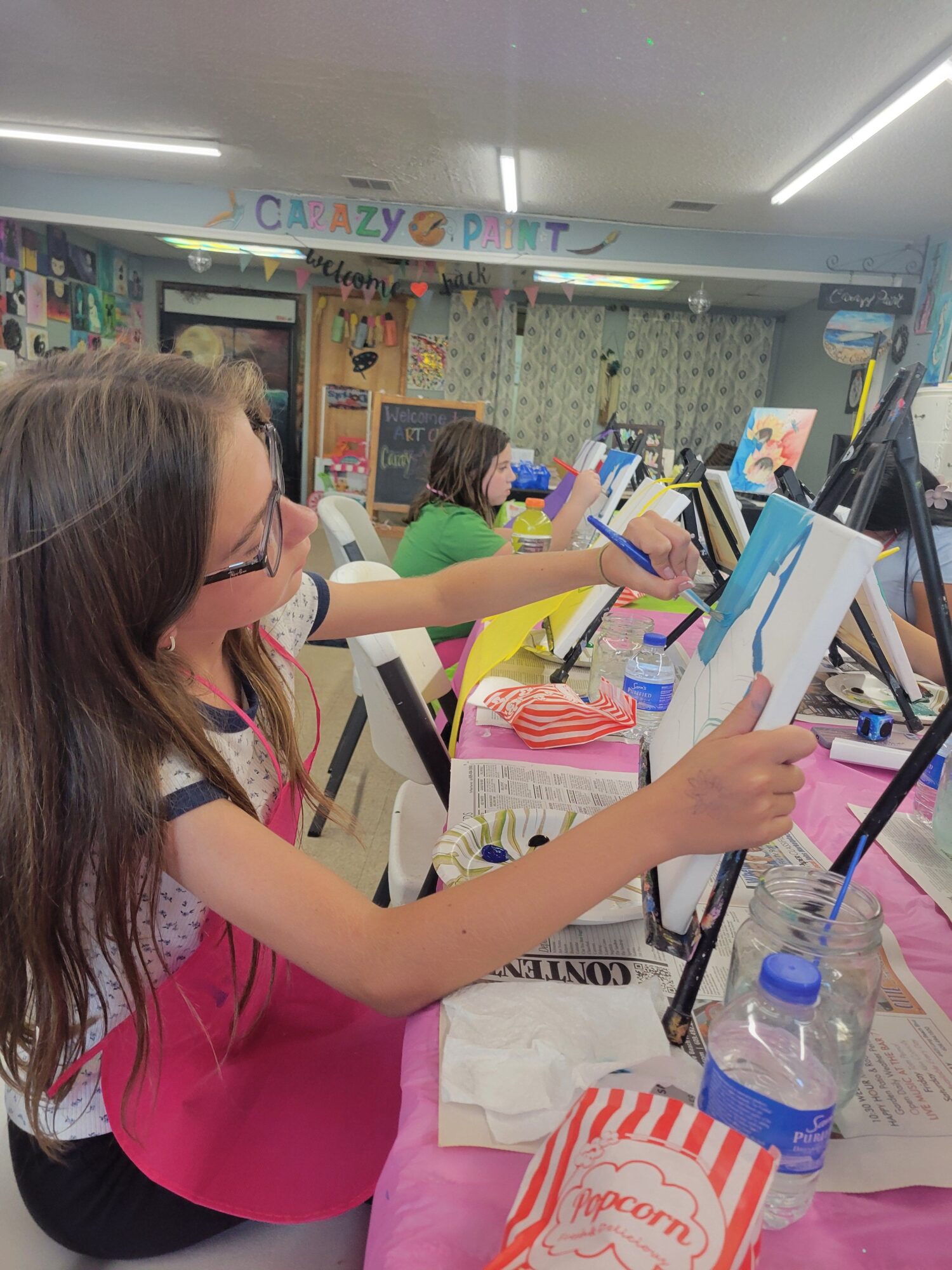 Girl with glasses and long hair writing at a table in a classroom, surrounded by books and supplies.