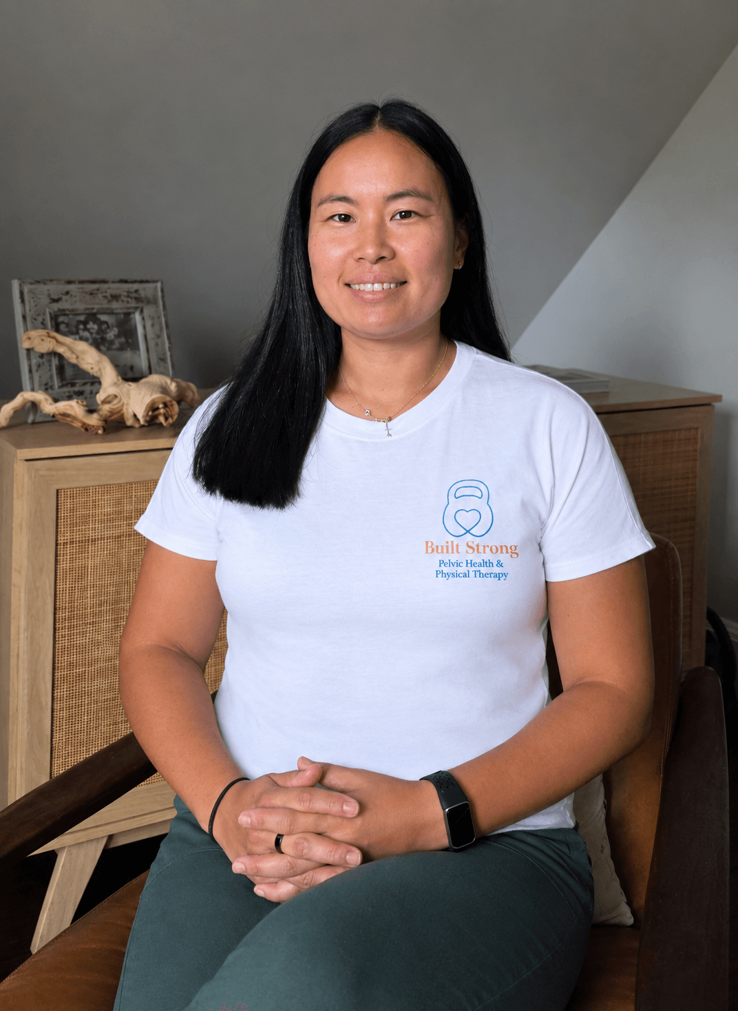 Woman with long black hair sitting indoors, smiling, wearing a white t-shirt and dark pants, with a wooden cabinet and decorative items behind her.