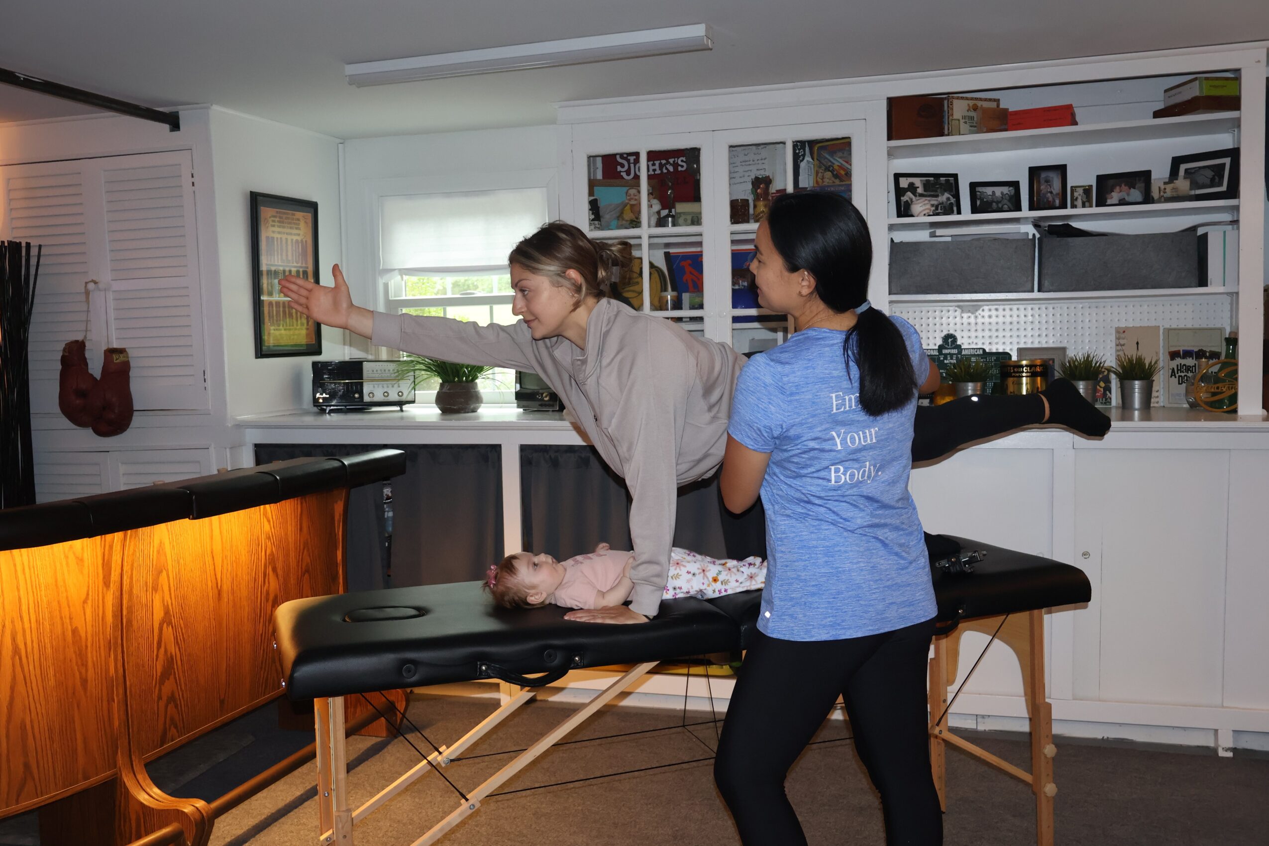 Two women in a room with shelves and a massage table, one woman reaching towards a television screen.
