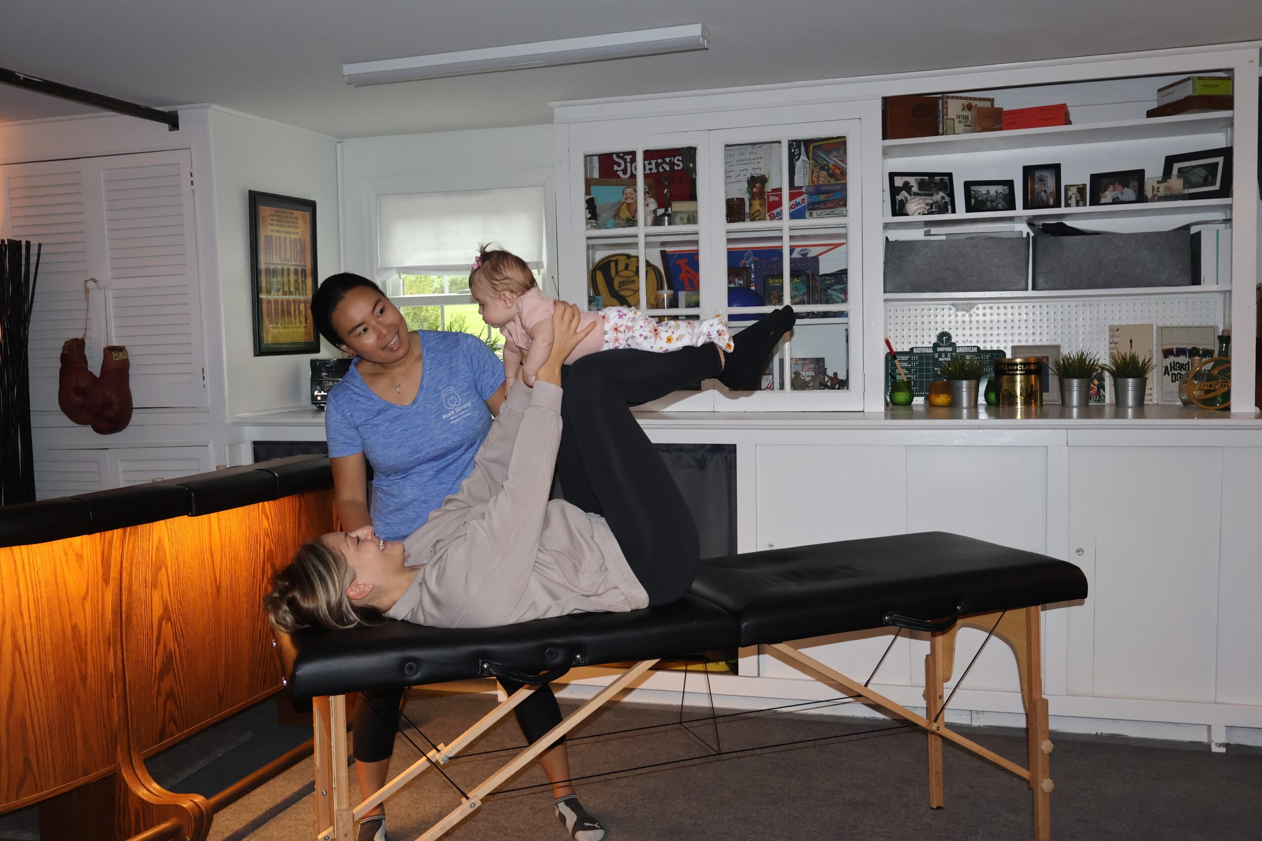 Two women and a child in a room with white shelves, one woman lying on a massage table, the other standing, holding the child.