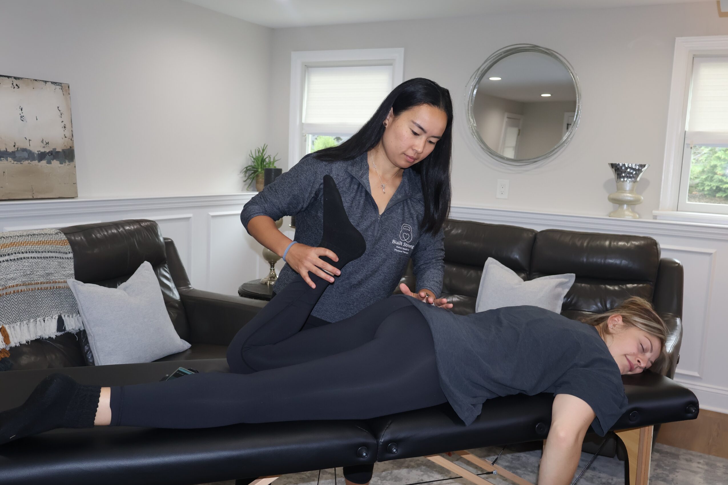 A woman receives a massage from a therapist lying face down on a massage table in a bright room with white walls and windows.