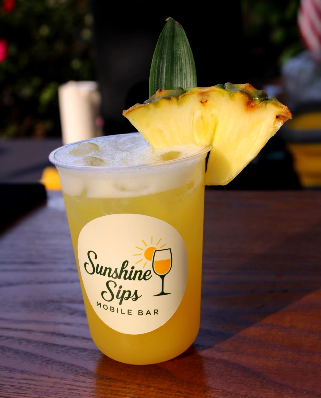 A pineapple drink with pineapple slice and pineapple leaf on a wooden table, with a logo reading 'Sunshine Sips Mobile Bar'.