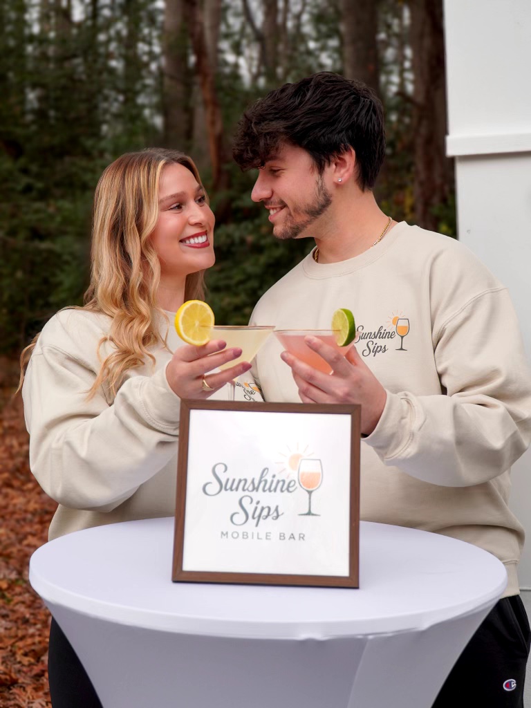Two people smiling and holding drinks with lemon slices, standing outdoors behind a table with a sign that reads 