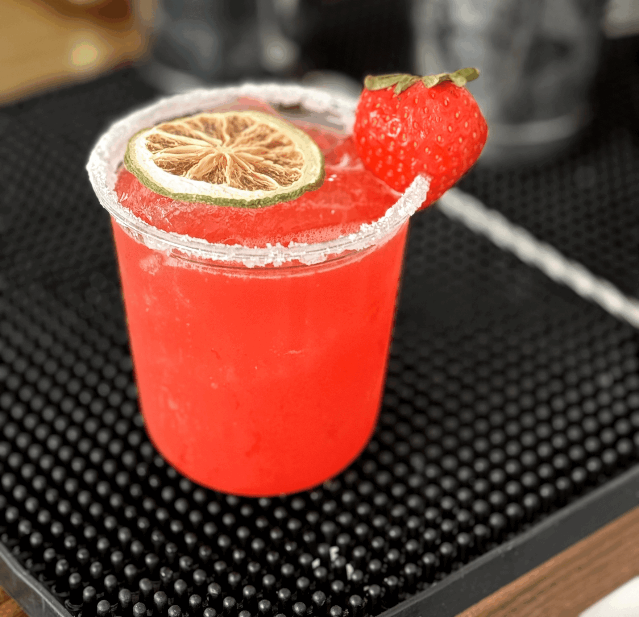 Red cocktail in a glass with a salted rim, garnished with a strawberry and a lime slice, on a black surface.