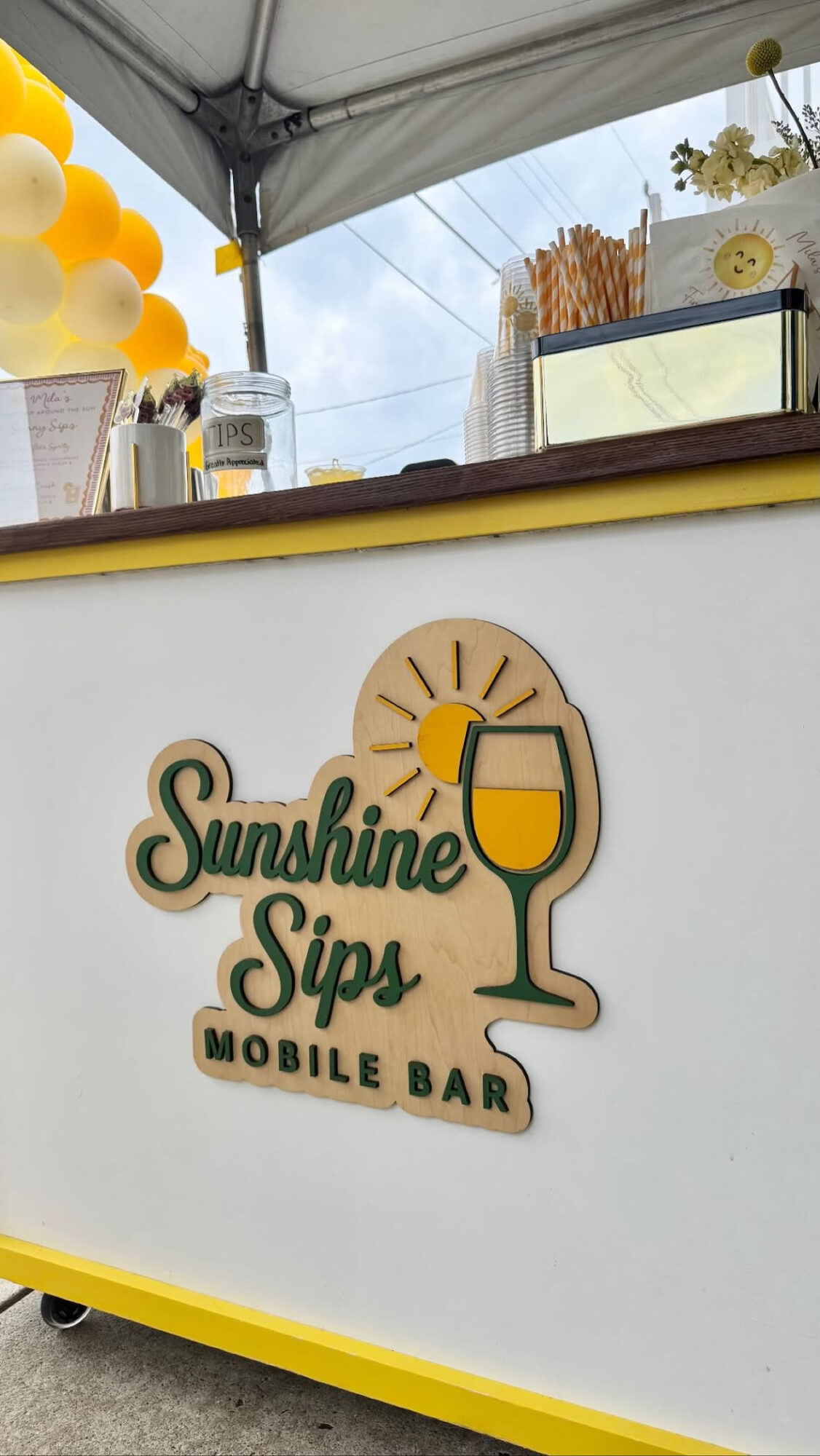 Mobile bar with a sign reading 'Sunshine Sips' and a glass of drink, decorated with yellow balloons.