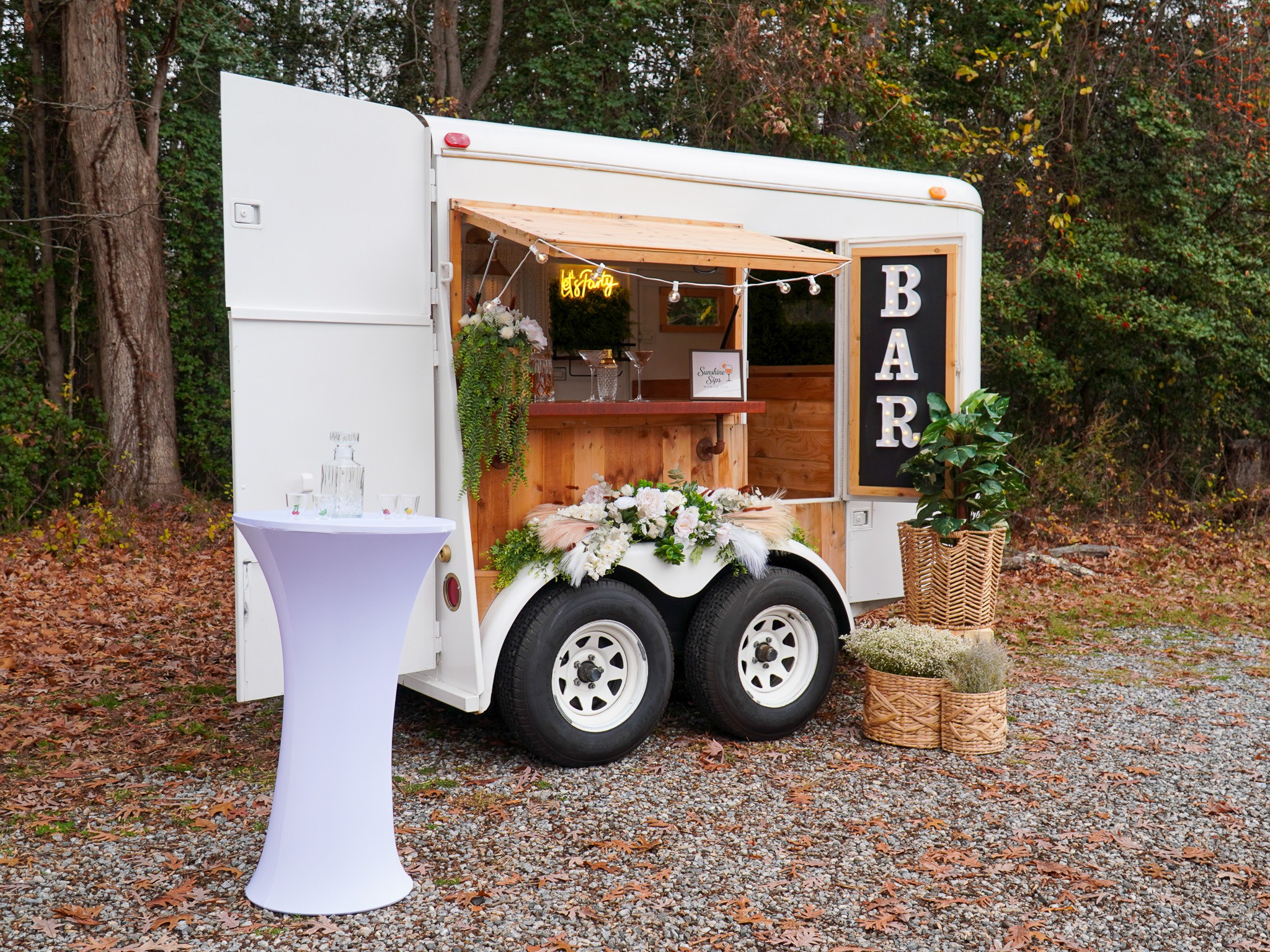 Food truck with bar sign, decorated with flowers and greenery, outdoors on gravel with trees in background.