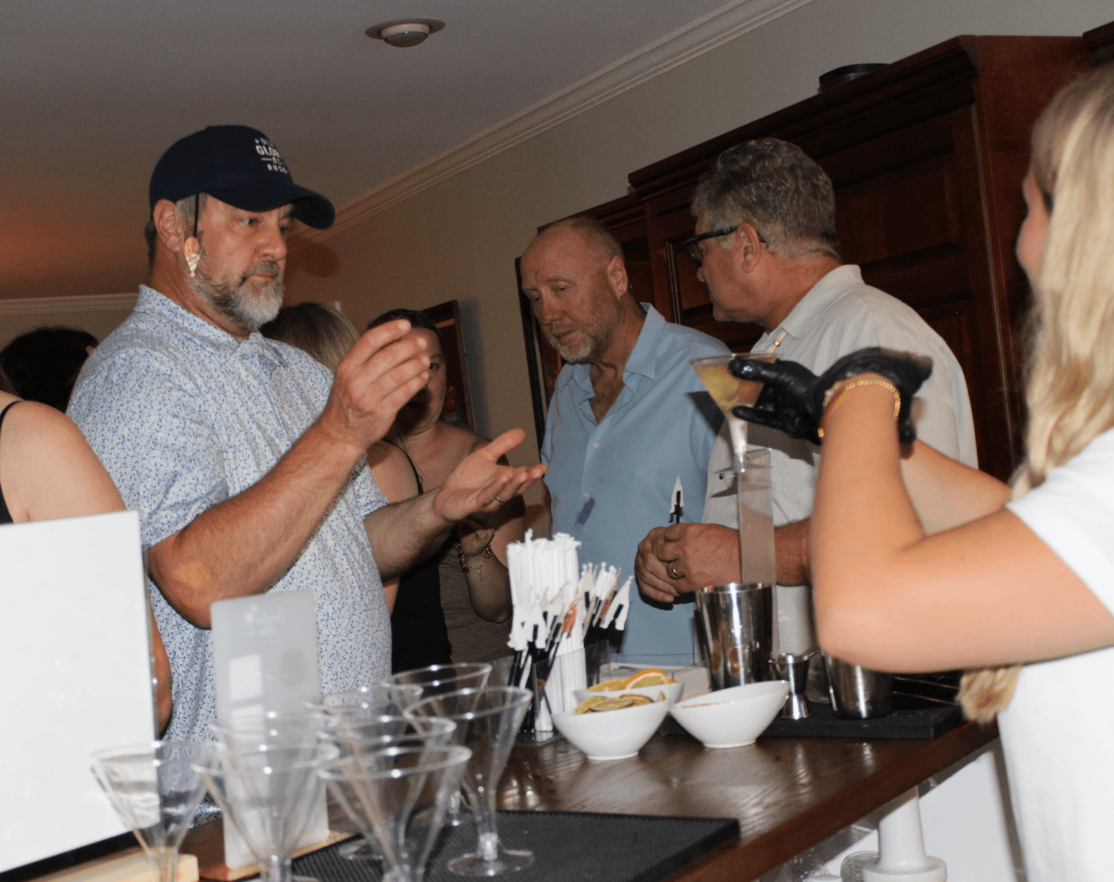 Group of people gathered around a bar, engaging in conversation and holding drinks, with barware and bowls on the counter.