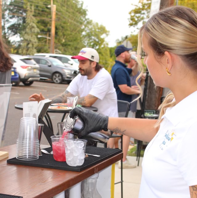 A woman pours a pink drink over ice at an outdoor event, with people and parked cars in the background.