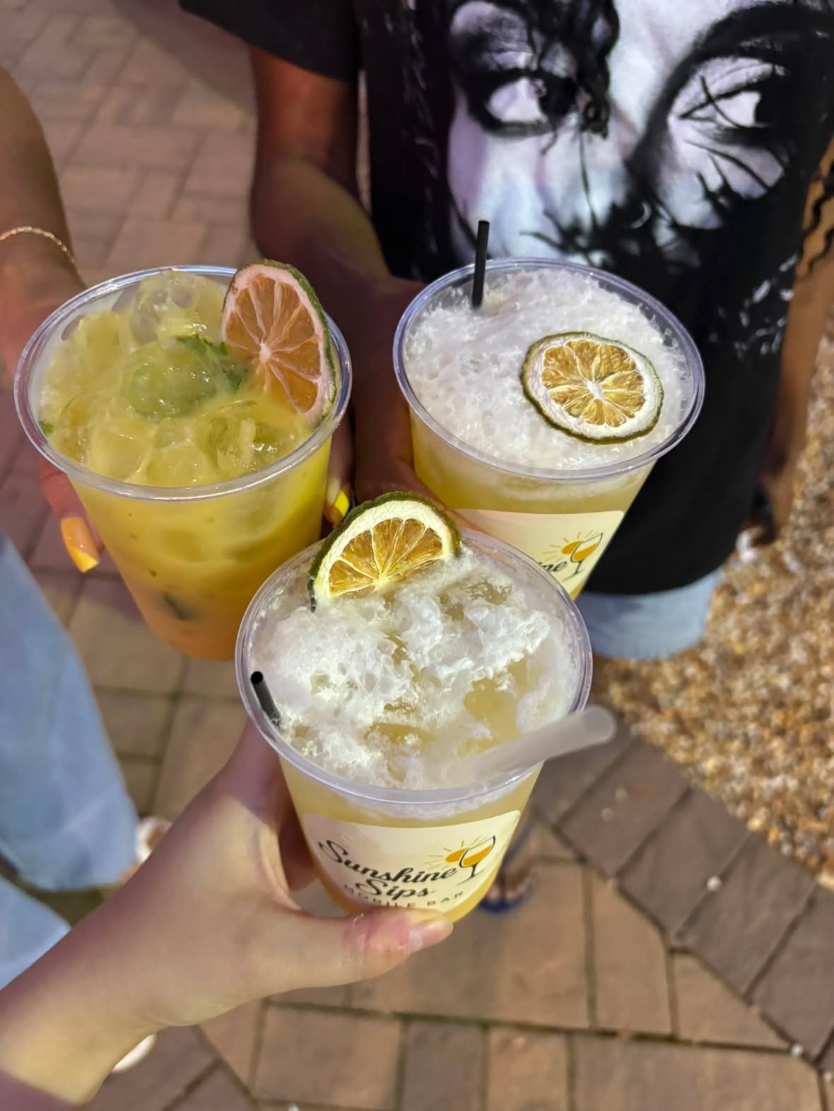 Three people holding drinks with lemon slices, one person wearing a black shirt with a face print, outdoor setting.