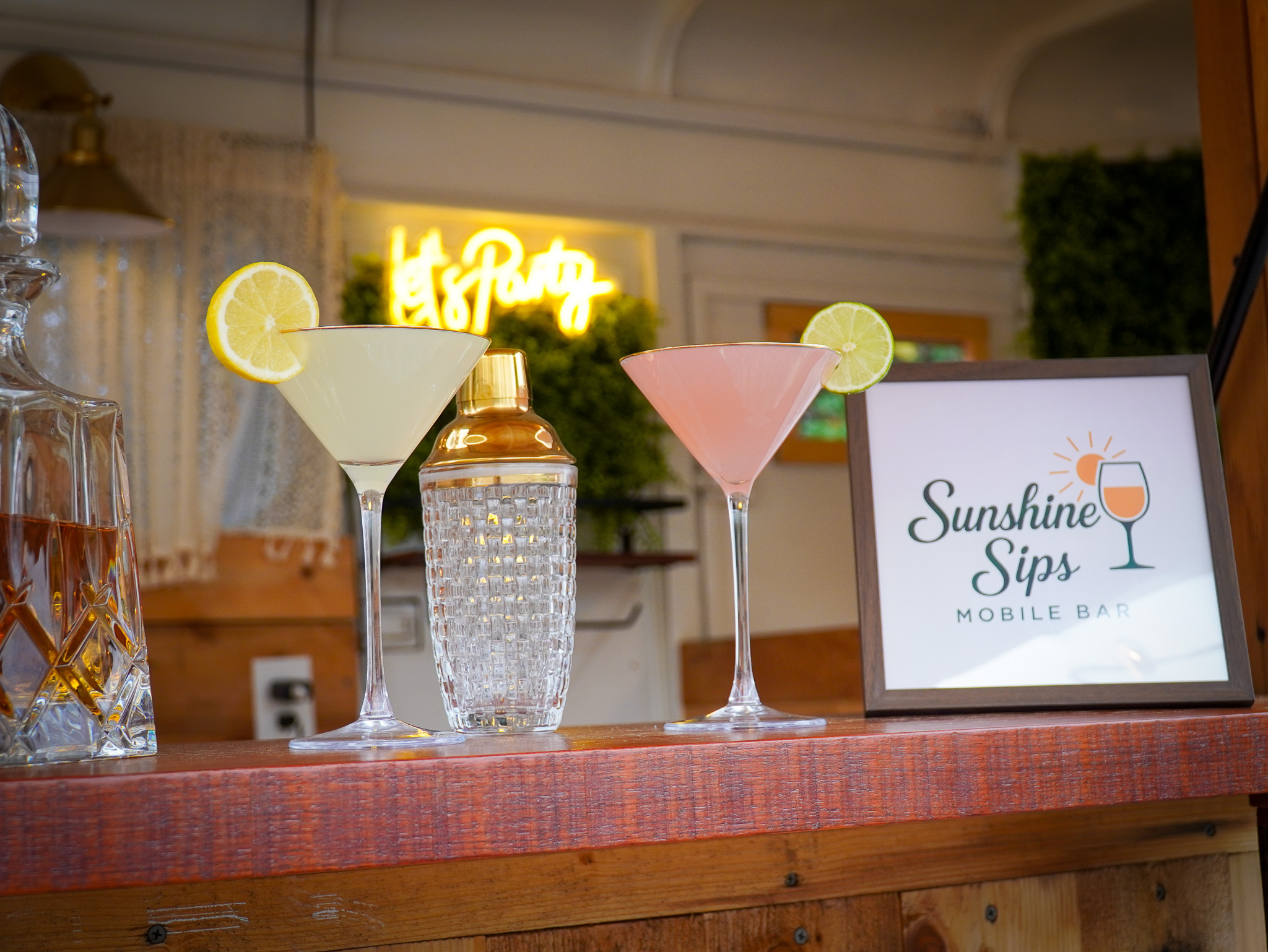 Three colorful cocktails with lemon garnishes on a bar counter, a framed sign reading 'Sunshine Sips,' and a neon sign in the background.