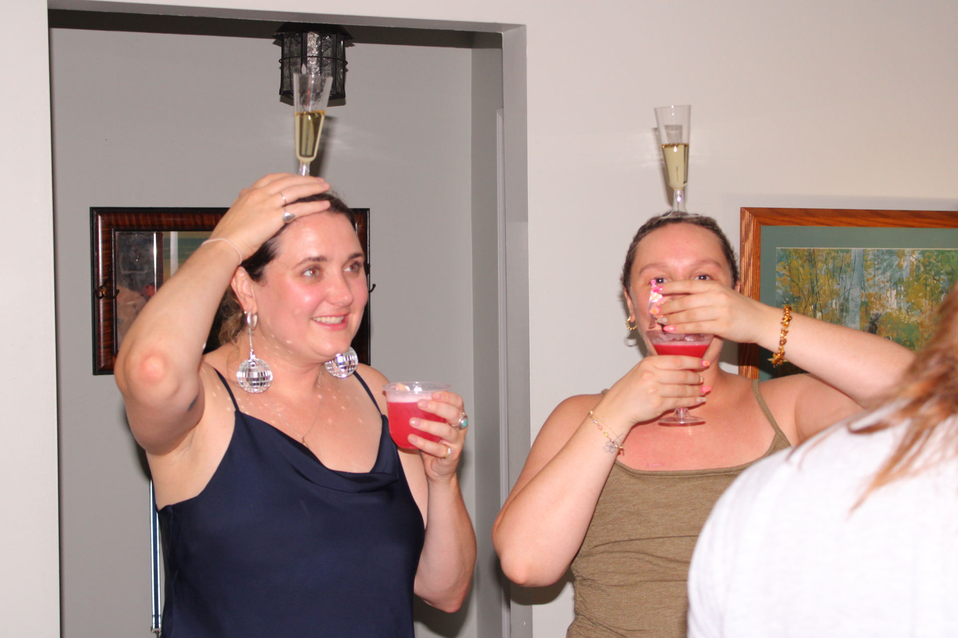 Two women holding drinks and smiling at a party, with a third person partially visible in the foreground.